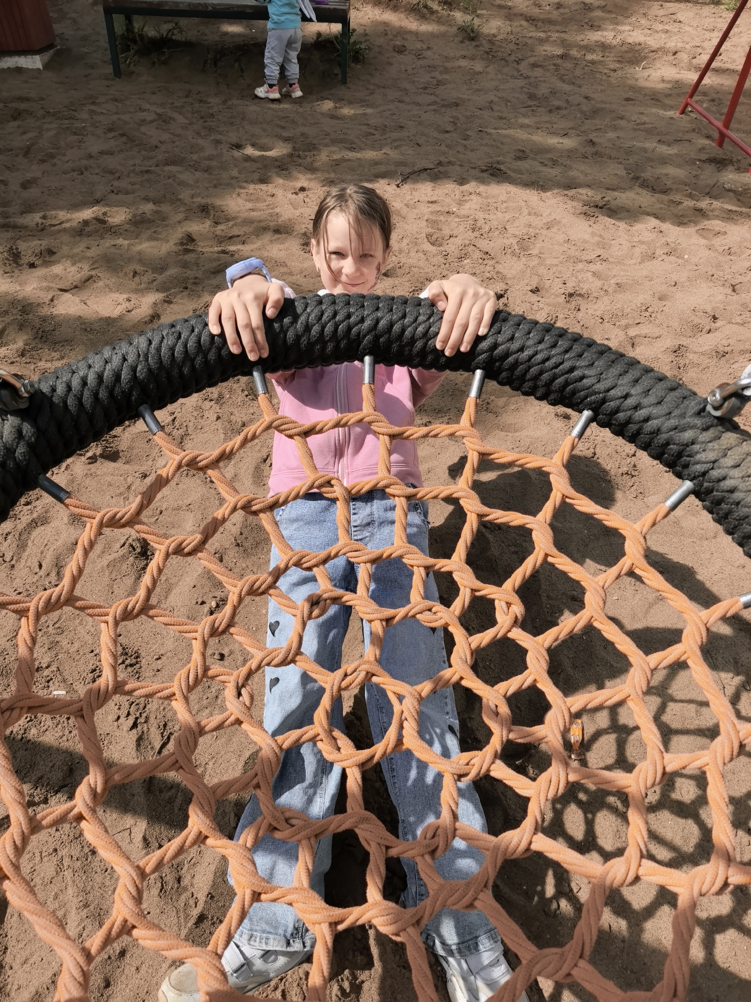 A young girl is depicted standing within a large circular rope swing, looking directly at the camera with a slight smile. She is centrally positioned in the frame, holding onto the thick black outer ring of the swing with both hands. The girl wears a pink zippered hoodie, light blue jeans adorned with black heart-shaped patches on the knees, and white sneakers. A blue wristband is visible on her left wrist, and there is a faint smudge on her right cheek.
The swing features a prominent orange knotted rope net, forming a web-like pattern that extends across the foreground and midground. The setting is an outdoor sandy playground, with the ground covered in light brown sand. Distinct shadows cast by the swing and the children suggest it is a sunny day, likely during late morning or early afternoon.
In the background, another child is partially visible, wearing a light blue top and grey pants, standing near a green wooden bench. A red metal structure, possibly part of other playground equipment, can also be seen further back. The overall atmosphere conveyed by the image is cheerful and playful. There is no visible text in the image.