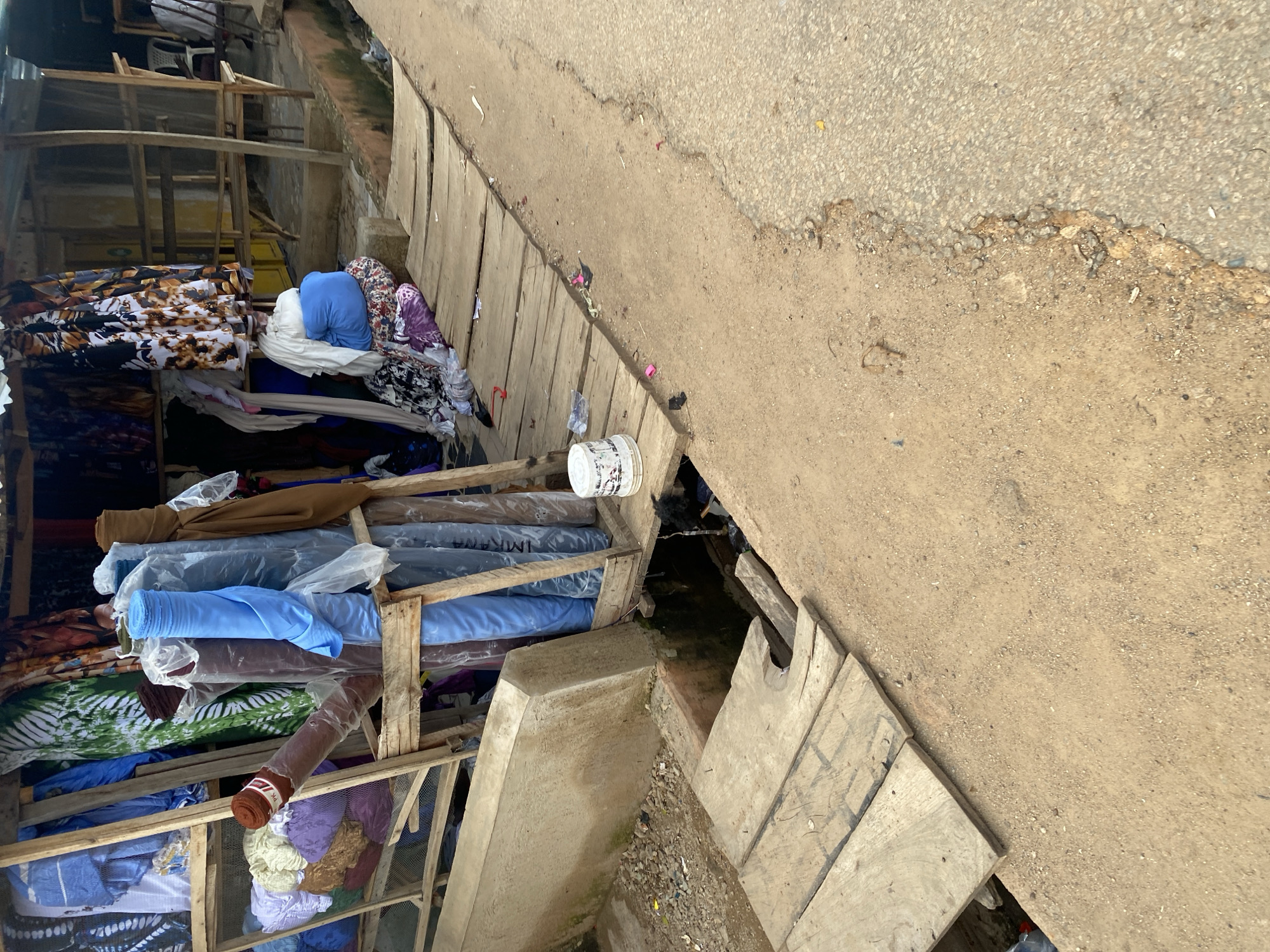 The image depicts an outdoor market stall, likely in Jos, Nigeria, specializing in fabrics. Several rolls of colorful fabric, predominantly in shades of blue, brown, and green, are stacked and displayed. Some are wrapped in plastic. A wooden structure serves as shelving for the fabrics. In the foreground, a rough wooden boardwalk extends over a ditch, which appears to be a drainage channel. The ground is a mix of dirt and gravel, with some scattered debris. A white bucket is placed on the boardwalk. In the background, more fabrics are visible, along with wooden supports for the market stall’s roof, which is made of corrugated metal. The overall impression is of a functional, if somewhat rustic, retail space in a bustling environment.
