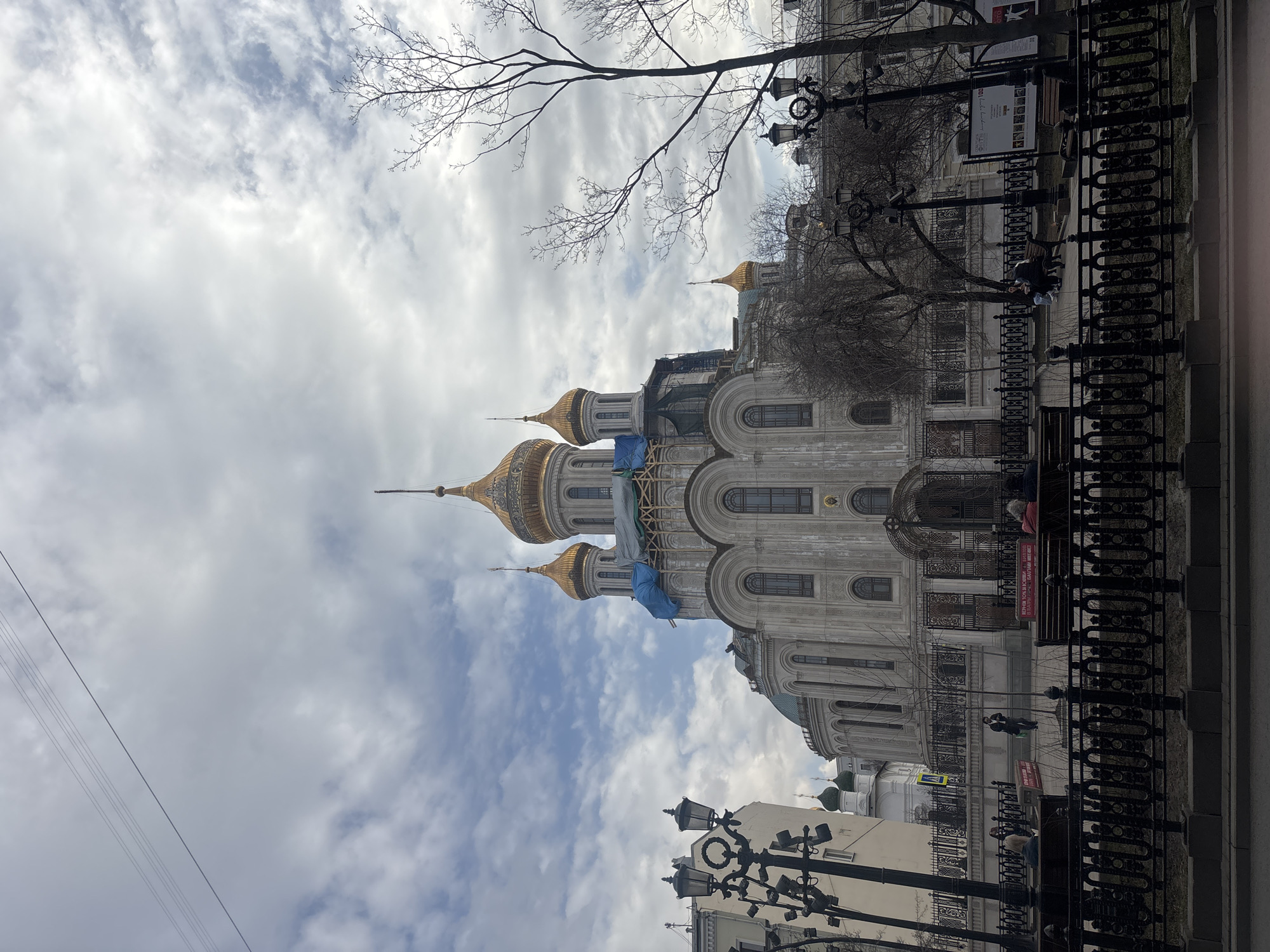 The image captures a view of the Sretensky Monastery in Moscow, Russia. The building features golden domes and is under renovation, with scaffolding and blue tarps visible. In front of the monastery, there is a decorative black fence and sidewalk where people are sitting on benches. Street lamps are interspersed along the sidewalk. The sky is cloudy, with patches of blue showing through. The photograph is taken at street level, looking up at the monastery. Bare branches of trees are visible in the background.