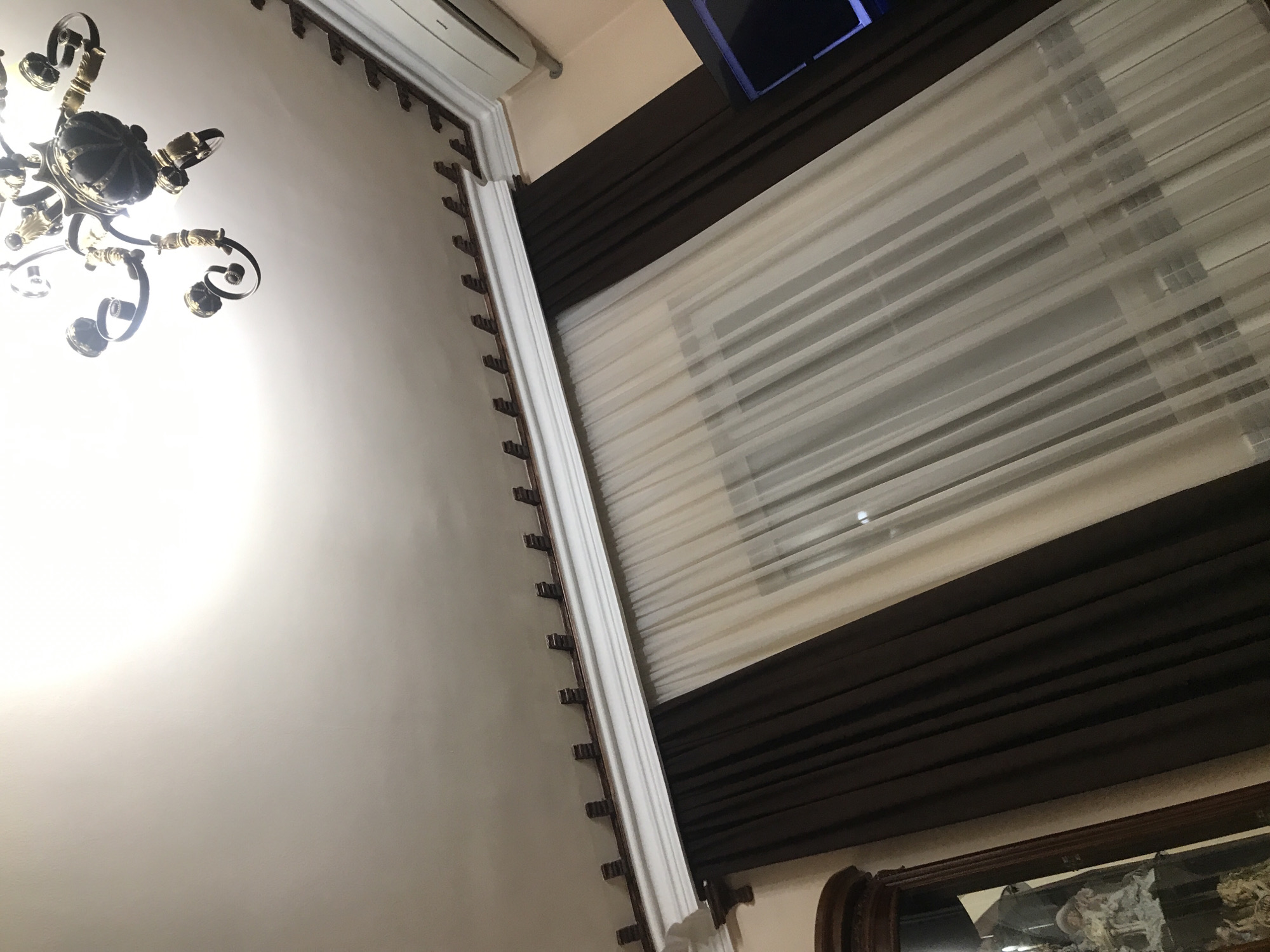 From a low-angle perspective, the image showcases a room with multiple architectural and decorative elements. The left side of the frame features a white wall illuminated by a chandelier with exposed bulbs. A decorative crown molding with regularly spaced pegs runs along the top of the wall.
On the right side, a window covered with a combination of sheer, horizontally striped blinds and brown curtains is visible. Above the window, part of a television screen and a white air conditioning unit is mounted on the wall.
At the bottom right corner of the frame, part of a dark-colored, ornate frame can be seen, suggesting the presence of a mirror or a painting.
The location is not explicitly identifiable as being in Yerevan, Armenia, without additional contextual information.