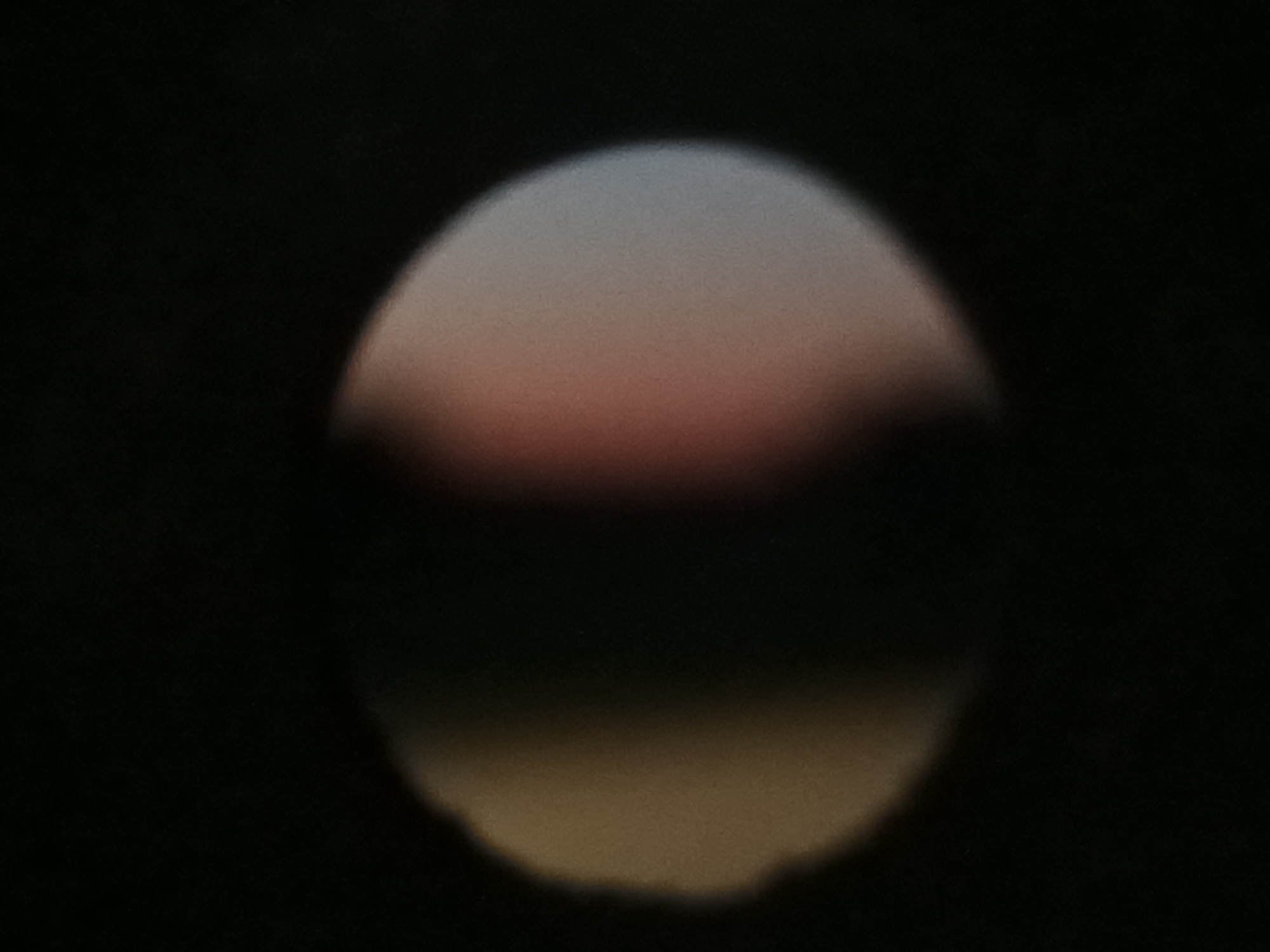 The image shows a blurry, circular depiction of a sunset or sunrise. The colors range from a pale sky blue at the top, transitioning through a soft pink and red, to a darker reddish-brown at the bottom. The background is predominantly black, suggesting nighttime or a very dark environment. There are no discernible people, objects, or specific environmental features that would indicate a particular location within Воскресенское, Russia. The overall impression is abstract and impressionistic, focusing on light and color rather than concrete details.