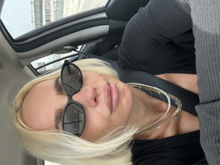 A fair-skinned female with short, blonde, bob-style hair and dark eyebrows is depicted in the front passenger or driver's seat of a vehicle. She wears black, narrow-frame sunglasses, under which her eyes are obscured and her nose shows a slight redness. Her full lips are coated with a light pink-nude gloss. Adorning her neck are multiple layered necklaces, including several gold-toned chains, some featuring small blue 'evil eye' charms, a thicker gold-toned chain, and two choker-style necklaces with small, clear stones (one gold-toned, one silver-toned). She is wearing a black ribbed long-sleeved top, partially unzipped to reveal cleavage, and is secured by a black seatbelt diagonally across her chest.
The vehicle's interior is light-colored, and the scene is illuminated by daylight. Through the passenger-side window and in the reflection of the left sunglass lens, an arid outdoor environment is visible. The reflection shows a modern multi-story building with a rectilinear, possibly glass, facade, adjacent to what appears to be an elevated road structure. Outside the vehicle, extensive ongoing construction is present, characterized by exposed sandy ground, scaffolding, concrete bridge abutments, and a prominent elevated highway or rail overpass in the mid-ground. This urban development and infrastructure, particularly the prominent highway structures and construction in an arid setting, are consistent with locations along major transportation corridors or rapidly developing areas within the city of Dubai, United Arab Emirates. The woman is looking directly towards the camera.