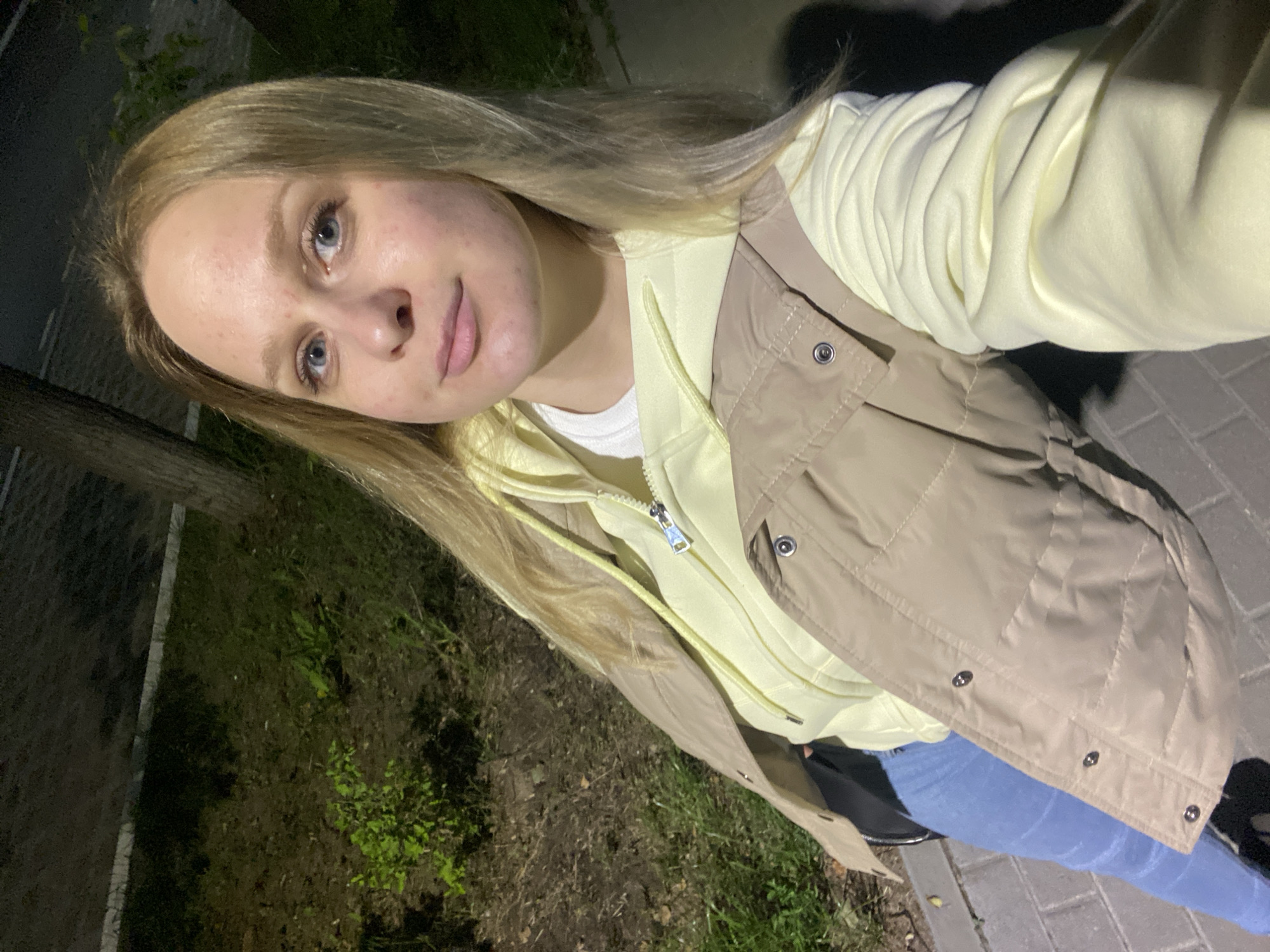 A young Caucasian woman with blonde hair and blue eyes is photographed from the chest up, in a selfie style, at night on a paved walkway in Chaikovsky, Russia. She is wearing a light yellow hoodie underneath a beige puffer vest. Her hair is long and straight, falling over her shoulders. The background consists of a grassy area with some trees and foliage, illuminated by artificial light.