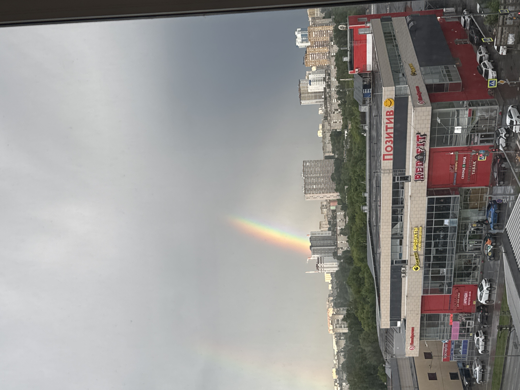 The image captures a view from an elevated position, likely a window, overlooking an urban landscape during a rainy day, punctuated by a rainbow. The sky is predominantly gray and overcast. A distinct rainbow arc is visible, stretching from the dark cloud cover towards a densely populated urban area. Buildings of varying heights and architectural styles, primarily residential apartments, are seen scattered among trees and green spaces.
The right side of the image features a commercial building complex. The building is multi-storied with a glass facade. There are signs such as "Позитив (Positive)", "RED FIT", "ОЧИЖИК ПРОДУКТЫ (Ochizhik Products)" and "Мастергаз (Mastergas)" visible on the building's exterior. The presence of cars and pedestrian crossings near the building indicates street-level activity.
The image seems to be taken in Perm, Russia, based on the Cyrillic lettering on the buildings and businesses depicted.