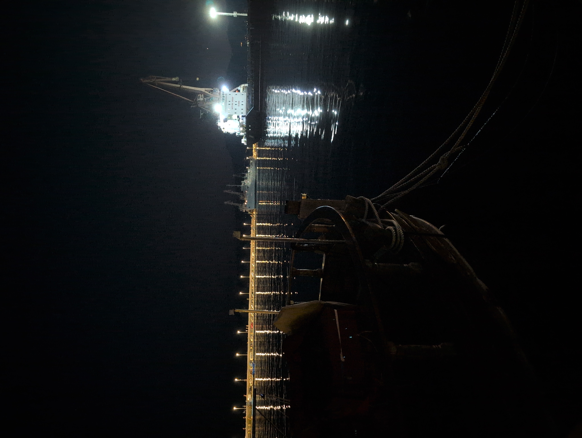 The image depicts a nighttime scene in a harbor. In the foreground, the dark silhouette of a boat is visible, with mooring ropes extending into the water. In the middle ground, a pier or breakwater is lined with streetlights, casting reflections on the calm water. In the background, a large cargo ship is illuminated by bright lights, with a crane positioned above it. Another ship is docked behind it. The scene appears to be located in the port of Rijeka, Croatia, a major maritime hub. The sky is dark and clear, suggesting a moonlit or star-filled night, though no celestial bodies are clearly visible. There are no people in the visible frame. The overall atmosphere is serene and industrial, highlighting the activity of the port even at night.