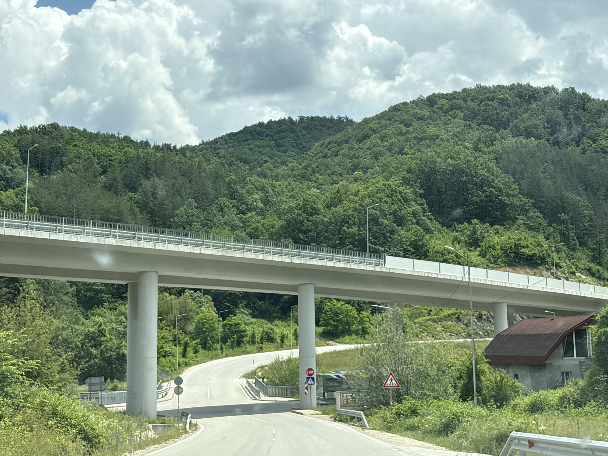 The image captures a section of a modern highway overpass constructed over a winding road in Gabrovo, Bulgaria. The overpass, supported by large concrete pillars, is a prominent feature, casting shadows on the road below. The surrounding environment is lush and green, with dense forests covering rolling hills that rise behind the infrastructure. The sky above is partly cloudy, with large white cumulus clouds against a blue backdrop.
On the road beneath the overpass, several traffic signs are visible. A red octagonal stop sign is positioned near one of the pillars, accompanied by a blue traffic sign depicting a pedestrian crossing. Further along the road, a triangular warning sign with a silhouette of a pedestrian is displayed. There is also a small, unconventional building with a reddish-brown, angled roof situated to the right of the road, partially obscured by foliage. The scene appears to be in a rural or semi-rural area, characterized by natural beauty and man-made infrastructure. No people are visible in the image.