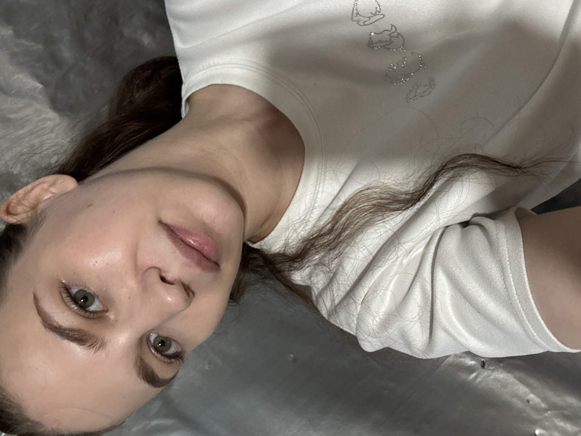A young woman with light brown hair and green eyes is positioned in the lower right of the frame, looking towards the camera. She is wearing a white t-shirt with a graphic design on the left side of the chest. Her skin appears smooth and slightly luminous. The background is a textured, silvery surface that appears to be a wall or curtain. The overall lighting is soft and diffused, creating a gentle glow on the subject's face. There are no visible indications of the location being in Nizhnyaya Gar', Russia.