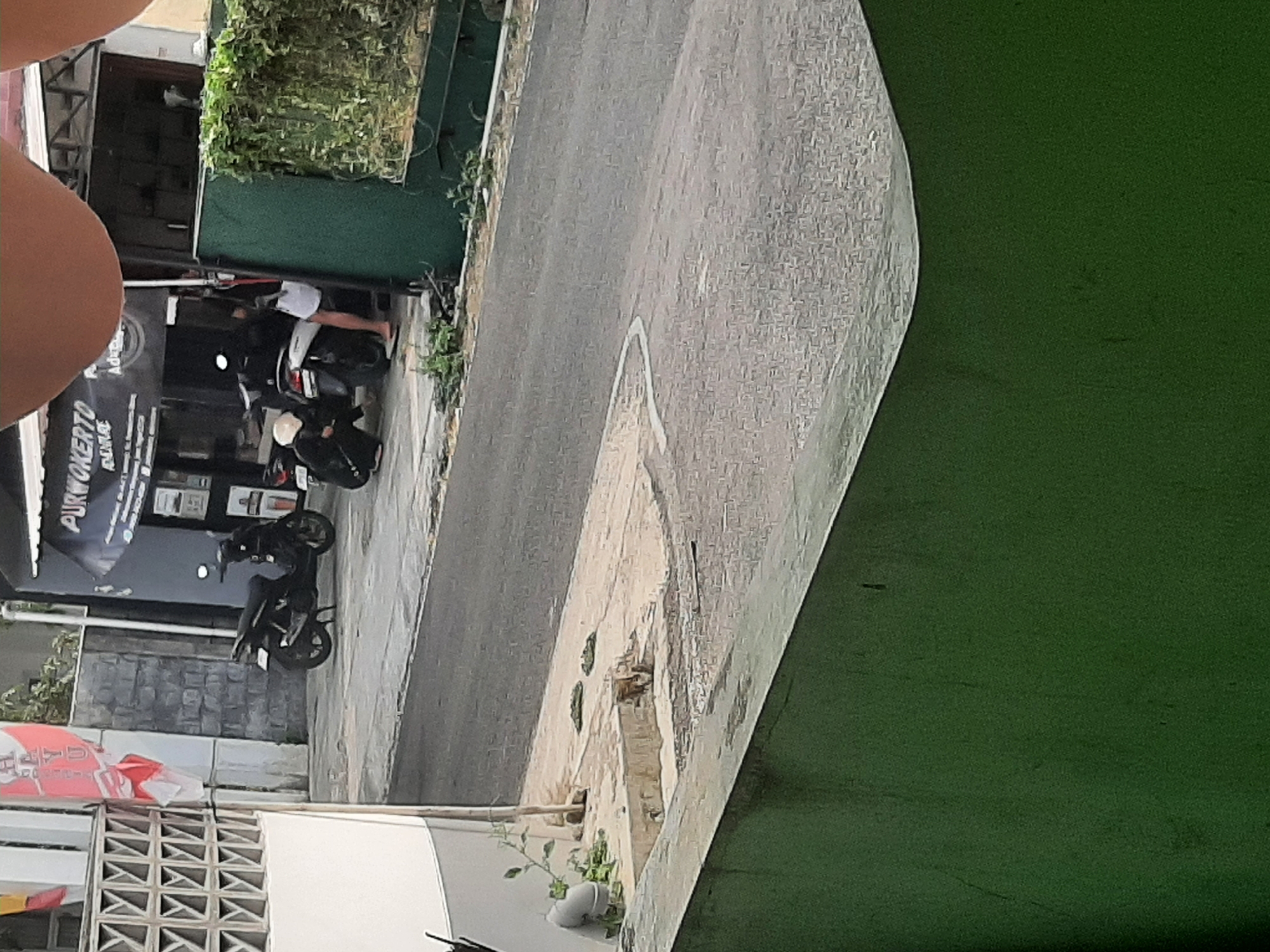The image depicts a street scene in Sumampir, Indonesia, likely an urban or semi-urban environment. In the foreground, a dark green wall dominates the lower portion of the frame, with a textured, lighter-colored surface above it. The middle ground features a paved road with visible markings, bordered by a concrete walkway. On the walkway, there are two parked motorcycles, one black and the other black with white accents. A person wearing dark clothing and a white helmet is crouched beside the white-accented motorcycle, appearing to interact with it. Another person, partially visible and wearing white shorts and flip-flops, stands near the black motorcycle.
Behind the motorcycles is the facade of a building with a dark awning and signage that reads "PURWOKERTO MAJU" and "ADVERT". A section of the building's exterior wall appears to be constructed of gray brick. To the left of the building, a white wall with a patterned ventilation grille is visible. Several small plants are growing in the vicinity of the walkway and the building. The background shows more of the street and the environment, including some greenery and the upper parts of buildings. The overall lighting suggests daytime.