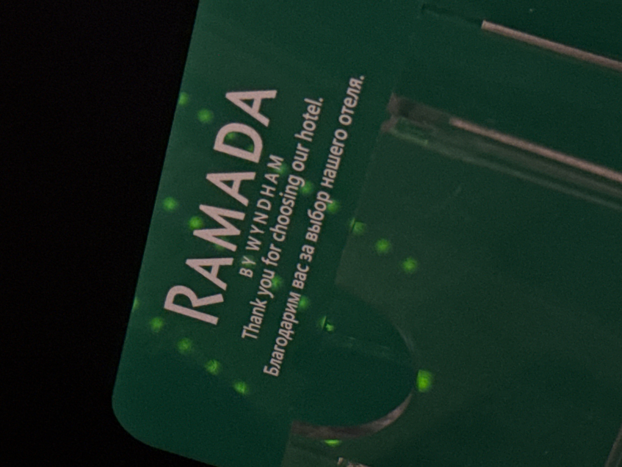 A green card with white text is displayed against a black background. The text reads "RAMADA BY WYNDHAM Thank you for choosing our hotel. Благодарим вас за выбор нашего отеля." There are several green spots, possibly reflections or lights, scattered across the card. The card is tilted, with the top left corner closer to the viewer. The bottom portion of the card is partially obscured by a clear plastic holder or stand. There are no people or animals visible in the image. The image is a close-up of a hotel amenity card, likely a welcome or thank you card from a Ramada hotel by Wyndham. The card is written in both English and Russian. The context of Nukus, Uzbekistan is not deducible from the image itself.