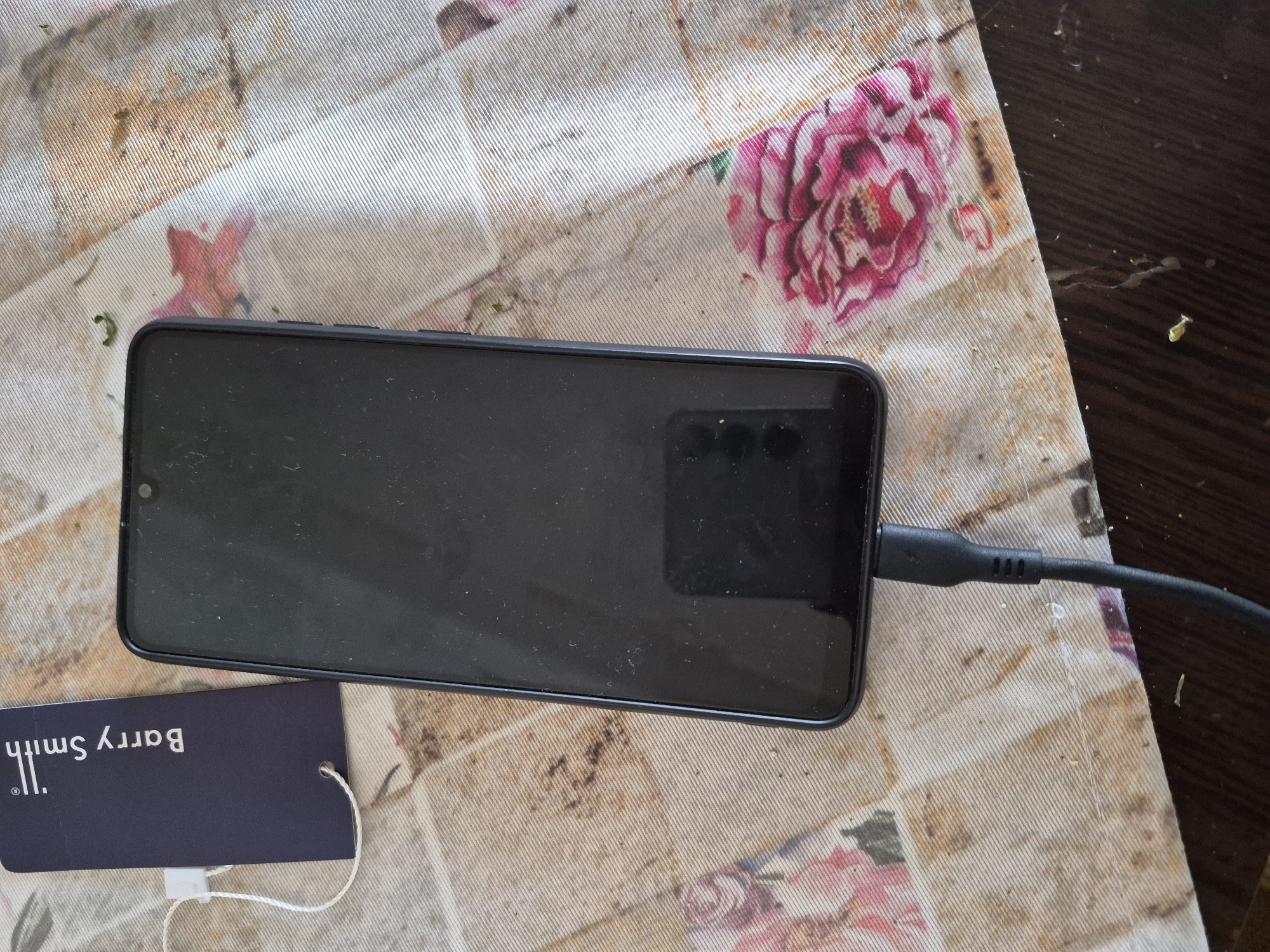 A black smartphone with a dusty, smudged screen and visible front camera notch lies on a patterned tablecloth. A black USB-C charging cable is plugged into the phone's right side, indicating it is connected for charging. To the left of the phone, a dark blue tag is present, displaying the upside-down text "Barry Smith," along with a registered trademark symbol and "II." A white string from the tag is also visible.
The tablecloth features a repeating floral pattern of pink and red roses on a light beige or white background. It shows signs of use, with multiple brown and green stains scattered across its surface, along with small specks of debris. Part of a dark brown wooden surface, possibly a table or floor, is visible to the right and top of the tablecloth.
The image captures an everyday indoor scene, likely in a domestic setting, as observed in Yerevan, Armenia. No individuals are present.