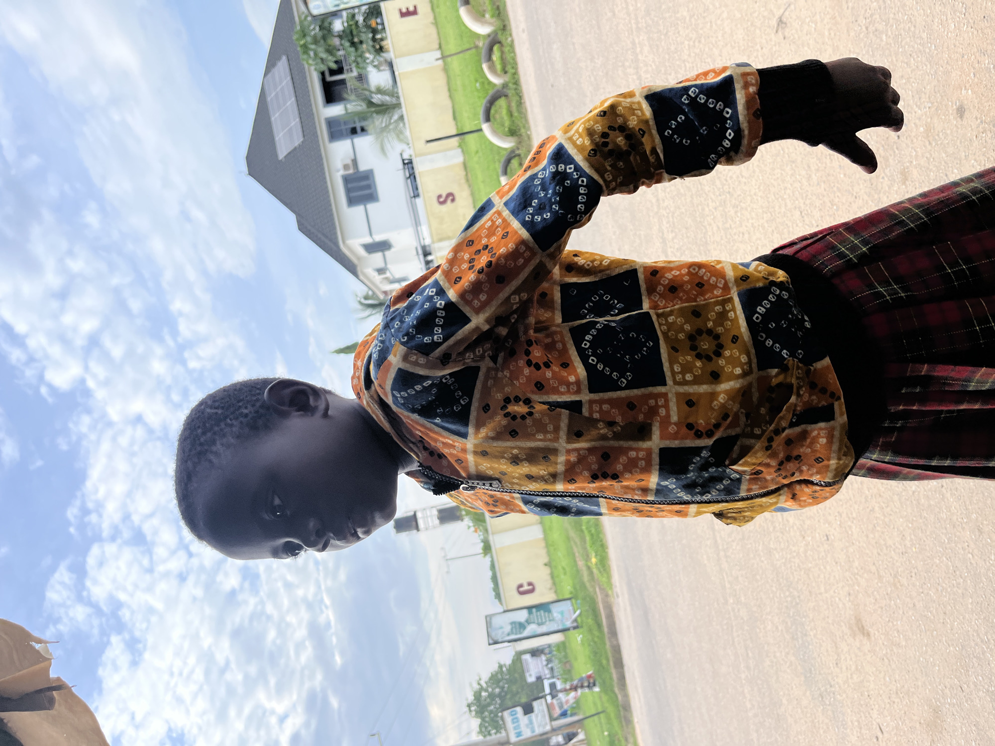 A young child, viewed from the chest up and positioned at an angle facing left, stands outdoors. The child is wearing a colorful, patterned zip-up jacket with squares of orange, blue, and yellow, featuring intricate white dotted patterns. A red and black plaid skirt is visible at the bottom of the frame. The child's left hand, covered in a black glove, is extended outwards. In the background, a modern white house with a dark roof and solar panels is visible. The environment appears to be a street in Abuja, Nigeria, indicated by the presence of signage with text, including "NSDC" and what appears to be "C" and "E" on fences, as well as streetlights and a water tower in the distance. The sky is partly cloudy with a bright, overcast condition.