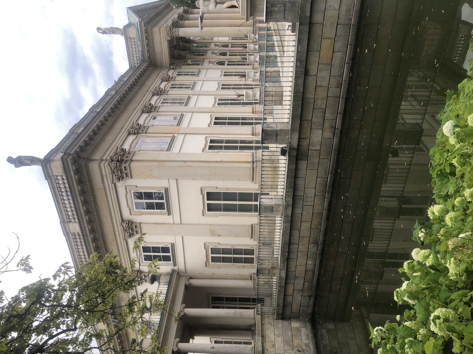 The image depicts the Palace on the Isle in Warsaw, Poland, a historic royal residence situated within the Łazienki Park. The palace, an elegant neoclassical structure, is flanked by a tranquil body of water. The water's surface reflects the building's facade, creating a symmetrical visual effect. Lush green foliage, including flowering hydrangeas, grows along the edge of the water, adding a touch of natural beauty to the scene. The sky above is overcast, with a mix of clouds. The palace's architecture is characterized by its classical columns, ornate detailing, and the presence of statues adorning its roofline. The overall scene conveys a sense of serenity and grandeur, highlighting the architectural splendor of the palace and its picturesque surroundings.
