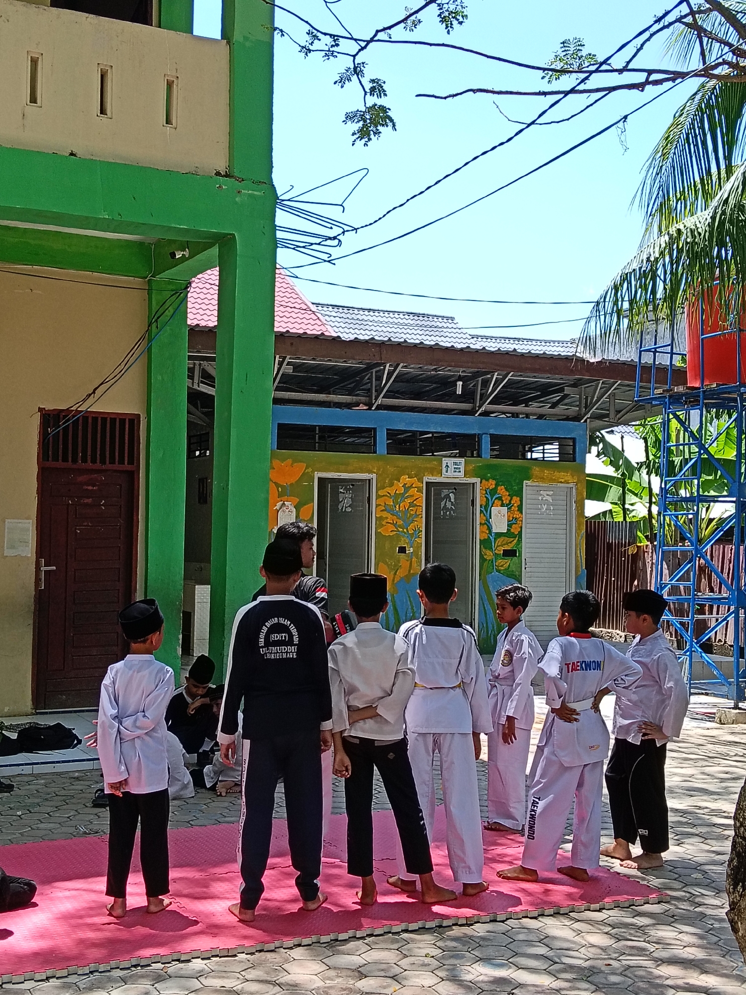 A group of young boys are gathered on a pink mat outdoors, possibly in Lhokseumawe, Indonesia, based on the text on clothing. Most of the boys are dressed in martial arts uniforms, some with "TAEKWONDO" visible on the back. One adult or older boy in a dark track suit stands with the group, facing the opposite direction. Another individual is seated on the mat. The backdrop includes a building with a green and cream facade and a brown door, along with a painted building designated as a toilet facility. Overhead are tree branches and power lines. The sky is clear and blue.
