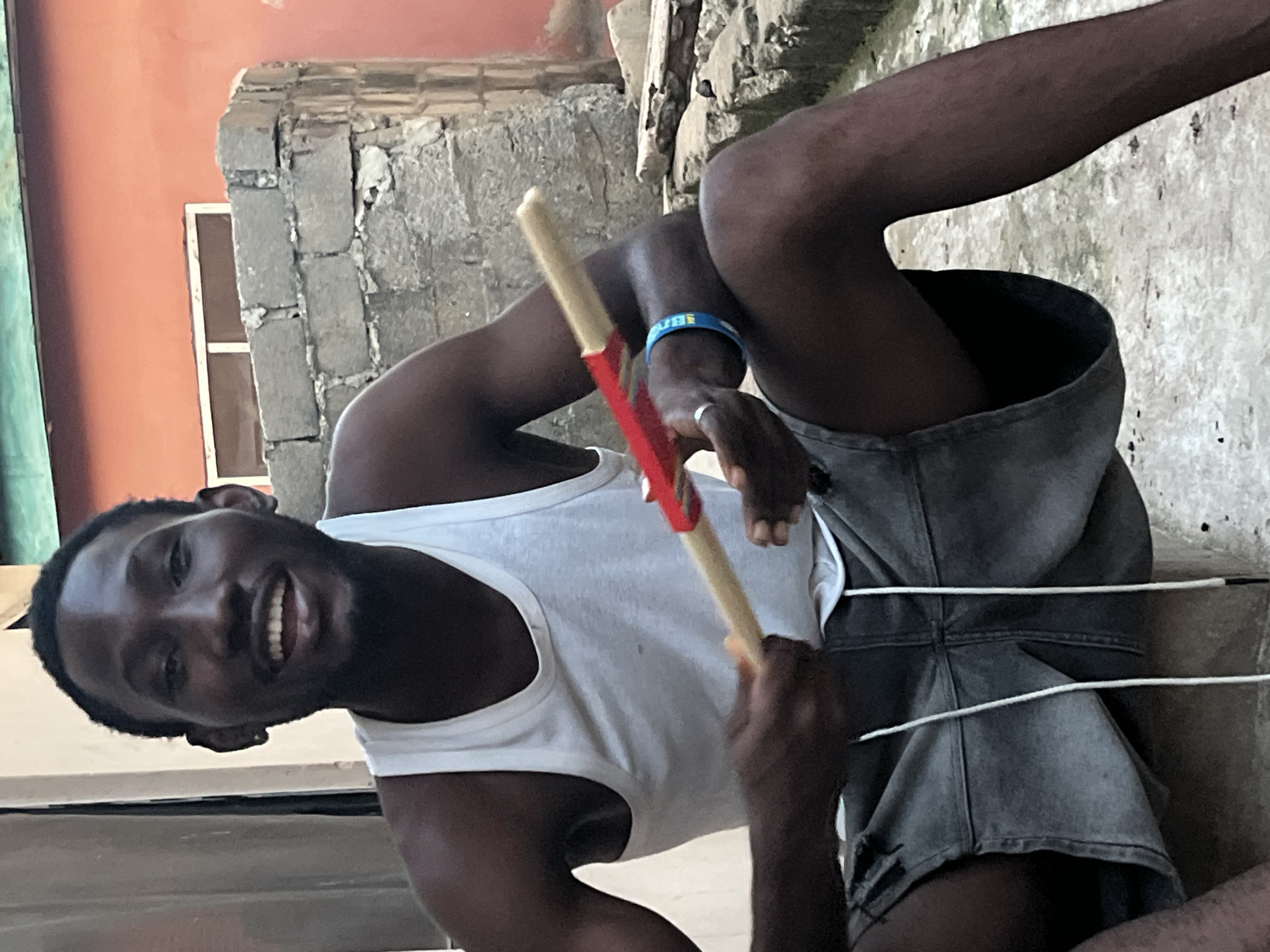 A man with dark skin and short black hair is sitting on a concrete step outdoors. He is wearing a white tank top and grey denim shorts. He is holding a long, cylindrical object with a red wrapping around the middle. He is smiling widely and looking at the camera. The background consists of a light-colored wall, a section of brick wall, and an orange-colored wall. There is also some rubble and vegetation in the background.
