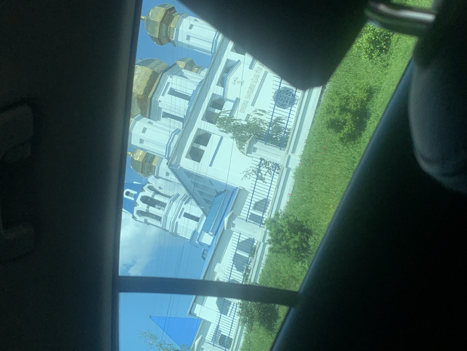The image shows a white building with blue accents, framed by a car window. The building, located at latitude 49.991, longitude 36.054 in Solonytsivka, Ukraine, appears to be a church or religious institution, featuring multiple gold domes and a sign that reads "Церква Зачаття Святого Івана Предтечі," which translates to "Church of the Conception of St. John the Baptist." The building is situated amidst lush green foliage. The camera is positioned inside a vehicle, looking out the window. The image captures the exterior of the building and some surrounding vegetation.