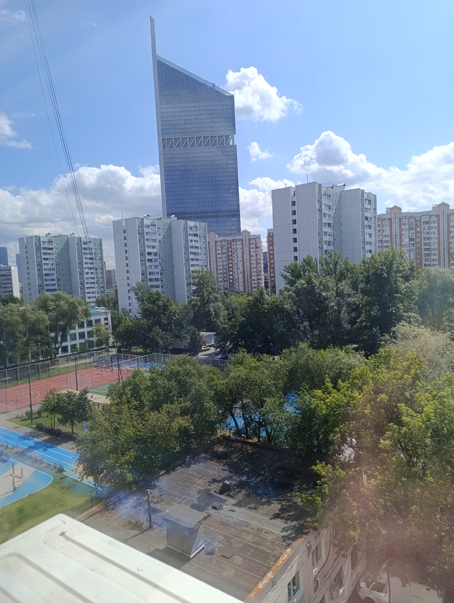 An aerial view shows a cityscape under a blue sky with scattered white clouds. In the background, a tall, modern skyscraper with a glass facade dominates the skyline. To its right are several multi-story apartment buildings. In the midground, a dense canopy of green trees obscures the view of more apartment buildings and a school building with light blue accents. A sports complex with a red and orange track and blue-lined courts is visible. In the foreground, the roof of a building is visible, with a ventilation shaft and a few scattered debris. A white van is parked next to a building on the right. The image is taken from a high vantage point, possibly from another building or a drone.
