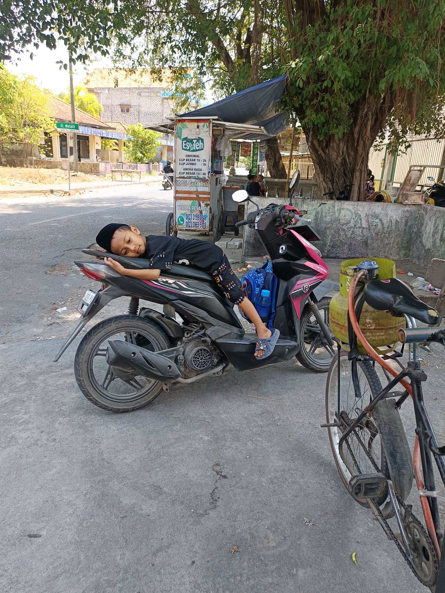 A young boy wearing a black skullcap and black clothing is sleeping on a black and pink motorcycle parked on an asphalt road. He is lying on his back with his head resting on the seat. A blue backpack with a water bottle is attached to the back of the motorcycle. In the foreground, the rear wheel of a bicycle with a green gas cylinder strapped to its frame is visible. In the background, a street vendor stall with a sign advertising "Ese Teh" (iced tea) is present. Buildings and trees line the street, and a street sign that reads "Jl. Melati" is also visible, indicating the location is Melati Street in Sampang, Indonesia. The scene appears to be a typical street view in a residential or commercial area.