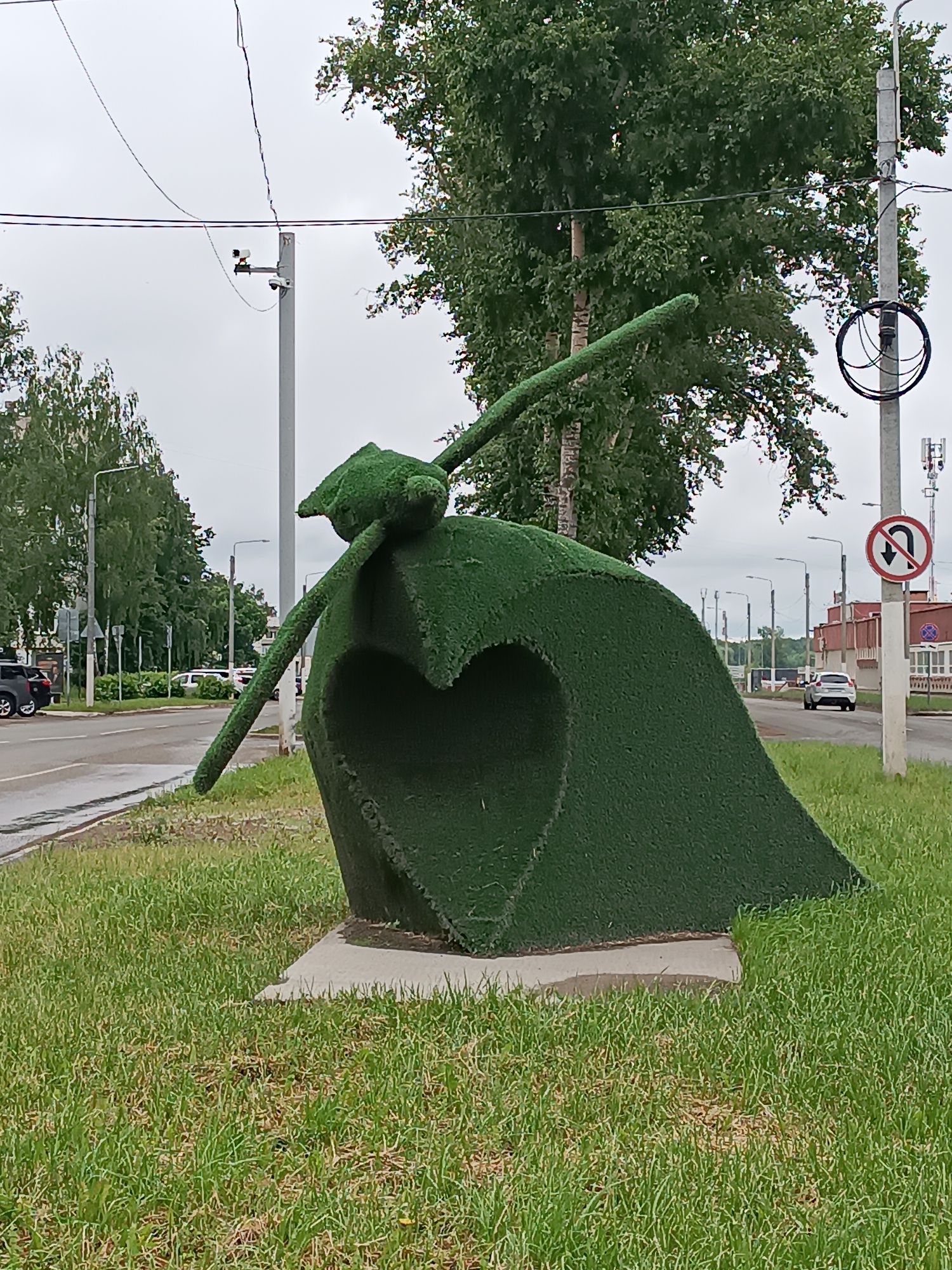 This image presents an outdoor urban scene in Chaikovsky, Russia, under an overcast sky. The central focus is an abstract sculpture covered entirely in artificial green grass, positioned on a strip of natural green grass adjacent to a paved road. The sculpture exhibits a large, hollowed-out heart shape on its lower section, with an elongated, upward-sloping element extending from its top. It is mounted on a light grey concrete base.
To the left of the sculpture, a multi-lane asphalt road with a broken white centerline shows signs of dampness. Several parked vehicles, including a dark SUV, are visible further down the road on the left. On the right side of the image, a white vehicle is seen moving away in the distance on the same road.
The environment is characterized by numerous tall, grey utility poles lining both sides of the road, supporting multiple strands of black electrical and telecommunication wires overhead. One utility pole on the left features a white camera device. Another prominent pole on the right side displays a circular red "No U-turn" road sign and is equipped with bundled black cables and cellular antennas. Lush green deciduous trees, including birch, are abundant, forming a verdant backdrop along the roadside. In the farther background on the right, several red-colored buildings with white window frames are discernible. The immediate ground is covered with natural green grass, interspersed with some drier patches.