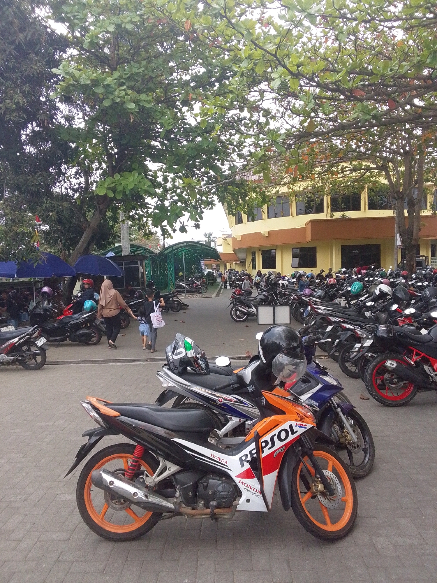 The image depicts a paved outdoor area in front of a two-story building with yellow and brown facades. The building has multiple dark-tinted windows. A canopy structure, supported by green metal beams, is visible in the middle ground, covering a section of the paved area. Numerous motorcycles are parked in rows, both under the canopy and in the open. Some motorcycles are in the foreground, with one notably featuring orange rims and "REPSOL" branding. In the mid-ground, a woman wearing a brown hijab and black pants is walking away from the viewer, holding the hand of a child wearing a blue shirt and shorts. Another child is walking beside them. Several blue umbrellas are set up in a shaded area, and various people are milling around in the background. Large trees with green leaves are prominent throughout the scene, casting shadows on the pavement. The overall atmosphere suggests a public space, possibly a parking lot near a commercial or educational establishment in Purworejo, Indonesia.