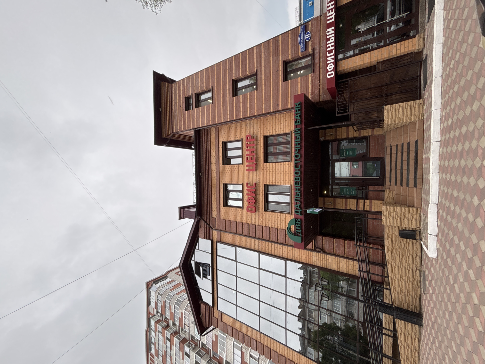 This image captures the exterior of a brick building with a glass facade, likely an office building or bank, located in Blagoveshchensk, Russia. The building features a prominent sign with Russian text, "ОФИС ЦЕНТР" (Office Center) and "ДАЛЬНЕВОСТОЧНЫЙ БАНК" (Far Eastern Bank). Another sign reads "ОФИСНЫЙ ЦЕНТР" (Office Center). A street sign indicates "Б. ХМЕЛЬНИЦКОГО" (B. Khmelnitsky), a street in Blagoveshchensk. The architecture includes a tall, narrow tower-like section. The surroundings show a patterned sidewalk and a glimpse of another building. The sky is overcast. There are no people visible in the image.