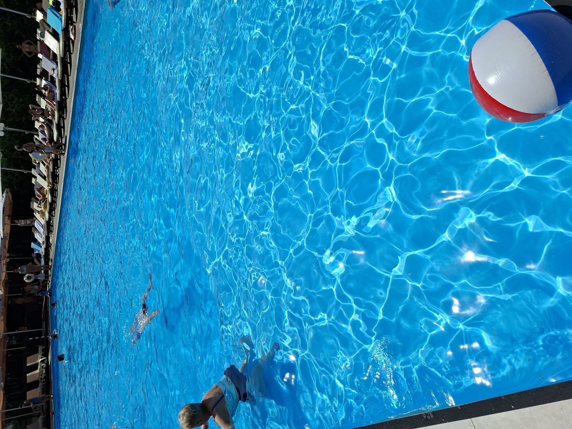 The image depicts a bright, sunny day at an outdoor swimming pool, likely located in Costești, Moldova. The pool's water is a vibrant blue, with sunlight creating shimmering reflections on its surface. In the foreground, a red, white, and blue beach ball floats near the edge of the pool.
Several people are visible in the water and around the pool. A woman with blonde hair is in the shallow end, facing away from the camera, appearing to be wading or standing in the water. Further out in the pool, a person is swimming underwater, with their body fully submerged and arms extended. More individuals are scattered throughout the pool, some with their heads above water.
On the far side of the pool, a row of lounge chairs is set up under large umbrellas, indicating a leisure area for sunbathers. Several people are seated or reclining on these chairs, enjoying the weather. The background shows a dense line of trees and vegetation, suggesting the pool is set in a natural or park-like environment. The overall scene conveys a relaxed and recreational atmosphere.