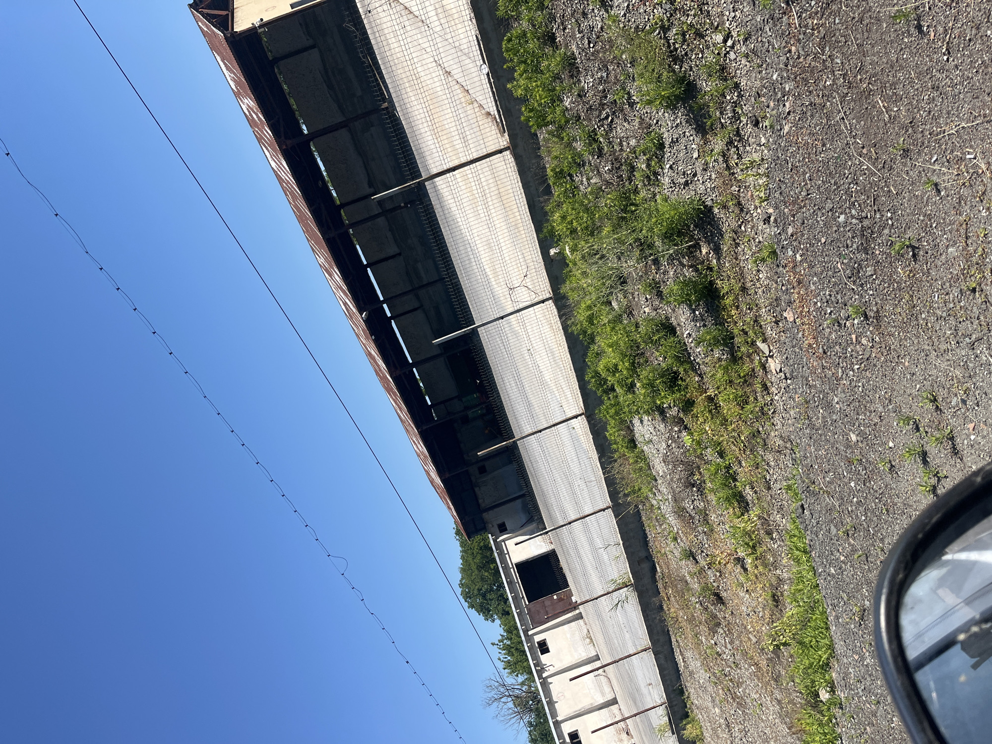 An outdoor, daytime, slightly angled medium shot shows a long, open-sided, warehouse-like structure with a rusty, corrugated metal roof and concrete foundation. The structure is situated in an area with sparse vegetation and gravelly ground, with a chain-link fence running along its length. To the left of the structure is a section of a white building with a large, open doorway. Above the structure, a clear blue sky is visible, with two parallel wires strung across it. The foreground of the image shows the edge of a car's rearview mirror, reflecting a portion of the scene. The location appears to be an industrial or storage area on the outskirts of Ravnets, Bulgaria.