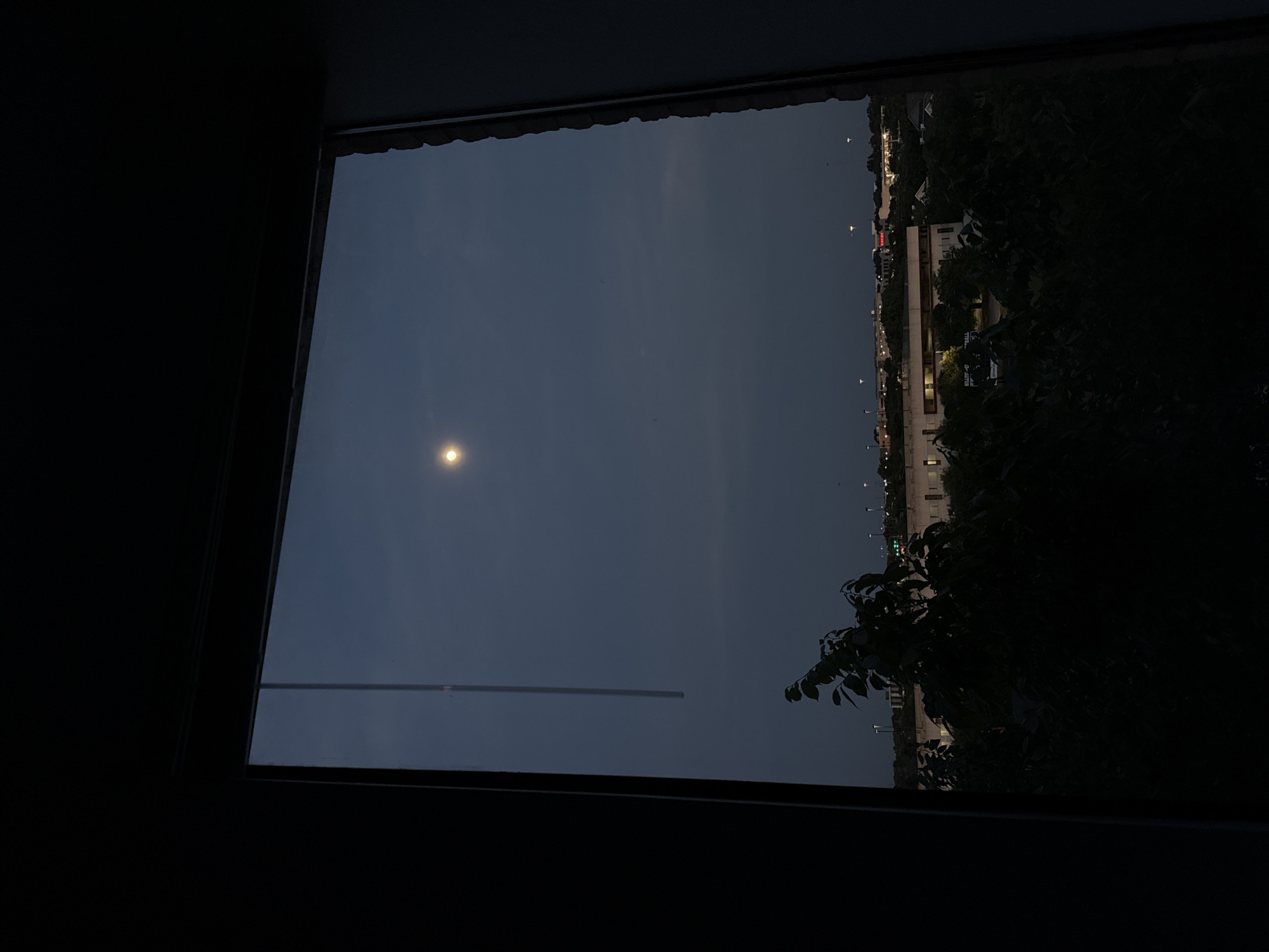 The image shows a view from a window in the city of Austin, Texas. The sky is dark, illuminated by a bright moon. A structure is visible through the window, as are other buildings with illuminated windows and street lights in the distance. Trees are visible at the side of the window.