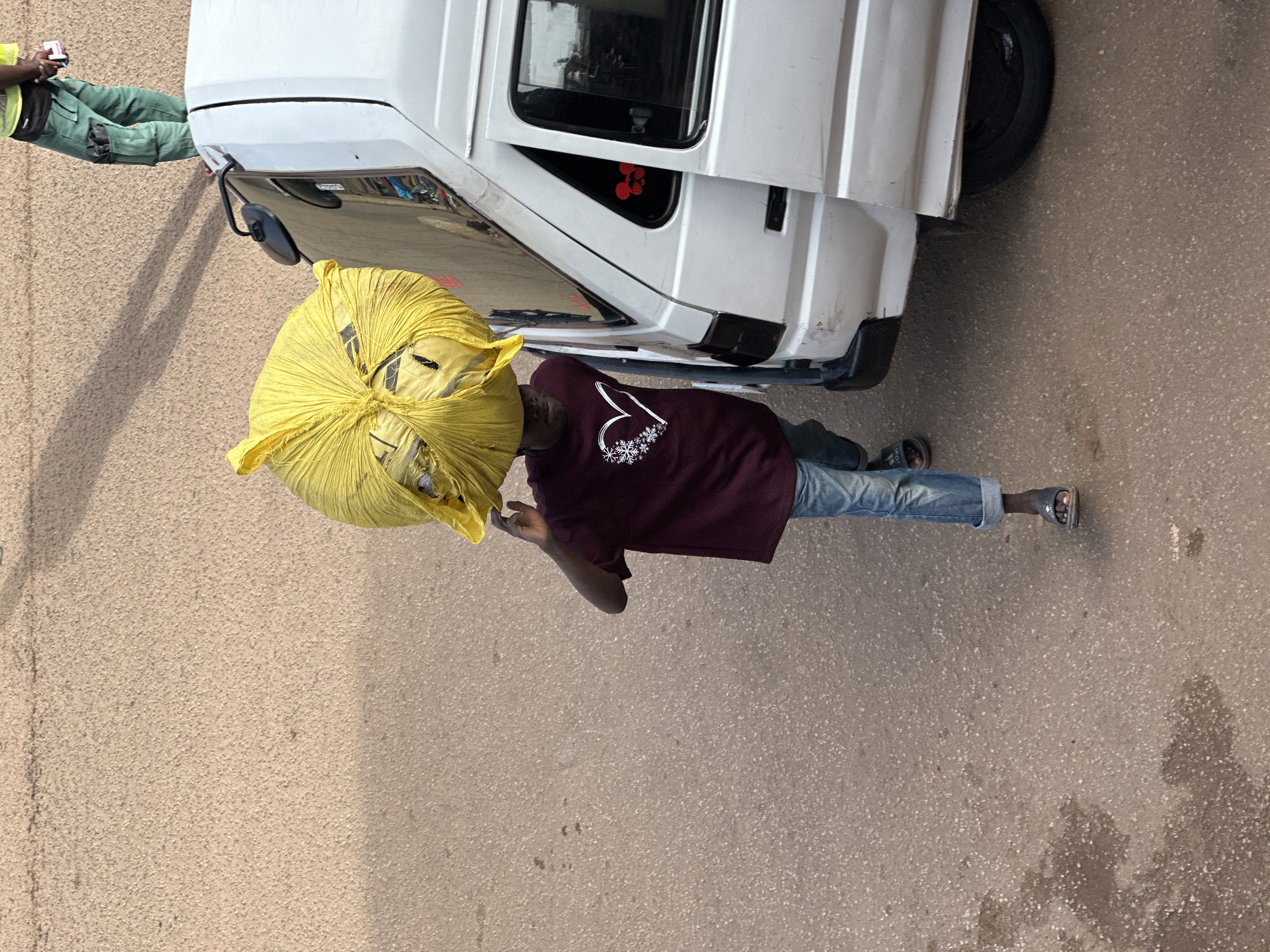 In Bukuru, Nigeria, a young person is captured from a high angle, balancing a large, bright yellow plastic bag on their head. They are walking on a dusty, asphalt road next to a white van. The individual is wearing a burgundy t-shirt with a white heart design and light blue jeans rolled up at the ankles. They are also wearing dark sandals. The van's doors are open, revealing the interior. Another person, dressed in green work pants and a high-visibility vest, stands near the van in the background, holding a small object. The scene suggests a moment of transport or delivery, with the environment being a typical urban street with scattered debris.