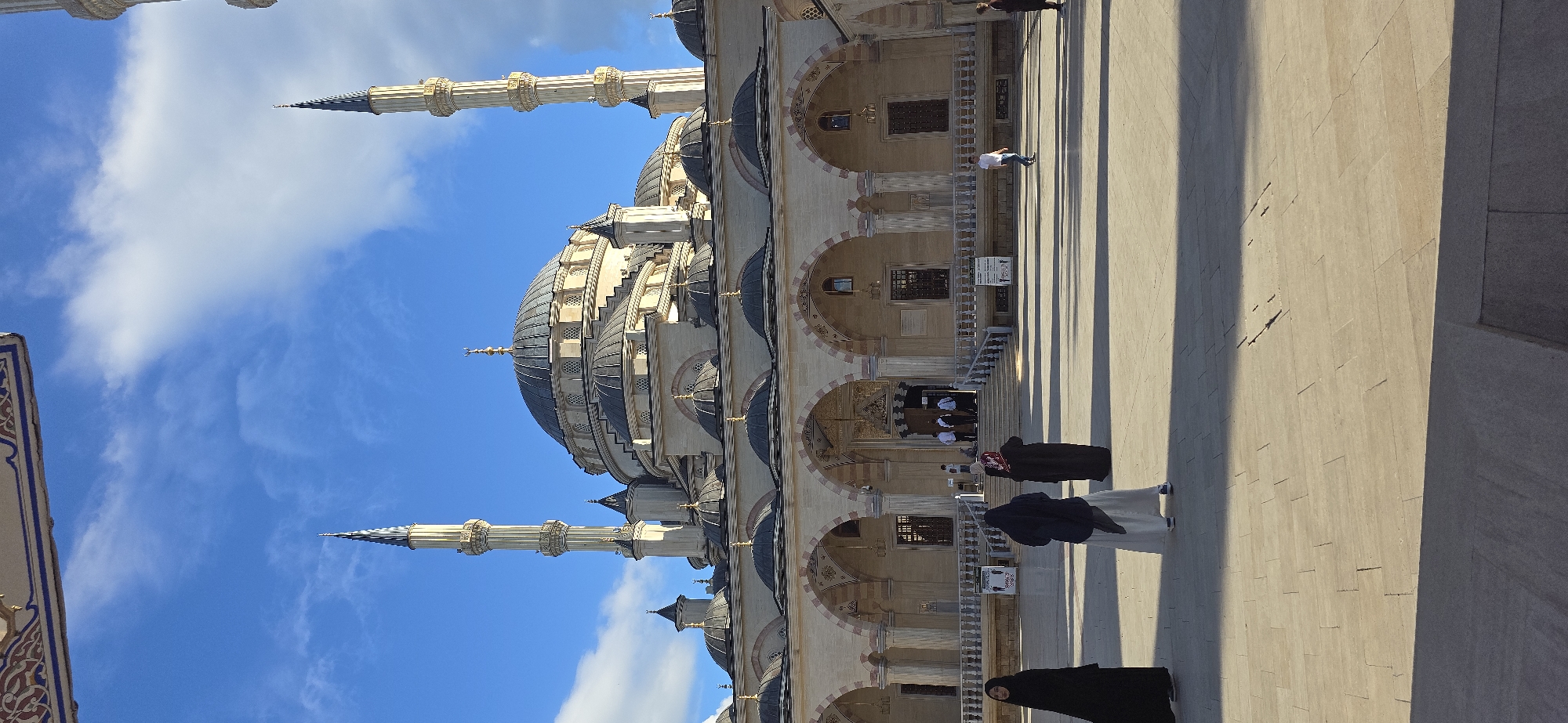The image shows the exterior of a mosque against a backdrop of blue sky with white clouds. The mosque features a large central dome and several smaller domes, with minarets extending upwards. The architectural style is ornate with arched windows and decorative details. In the foreground, several people are walking, some wearing traditional Muslim attire. The location is likely in the city of Grozny, Russia. The environment is sunny and bright.