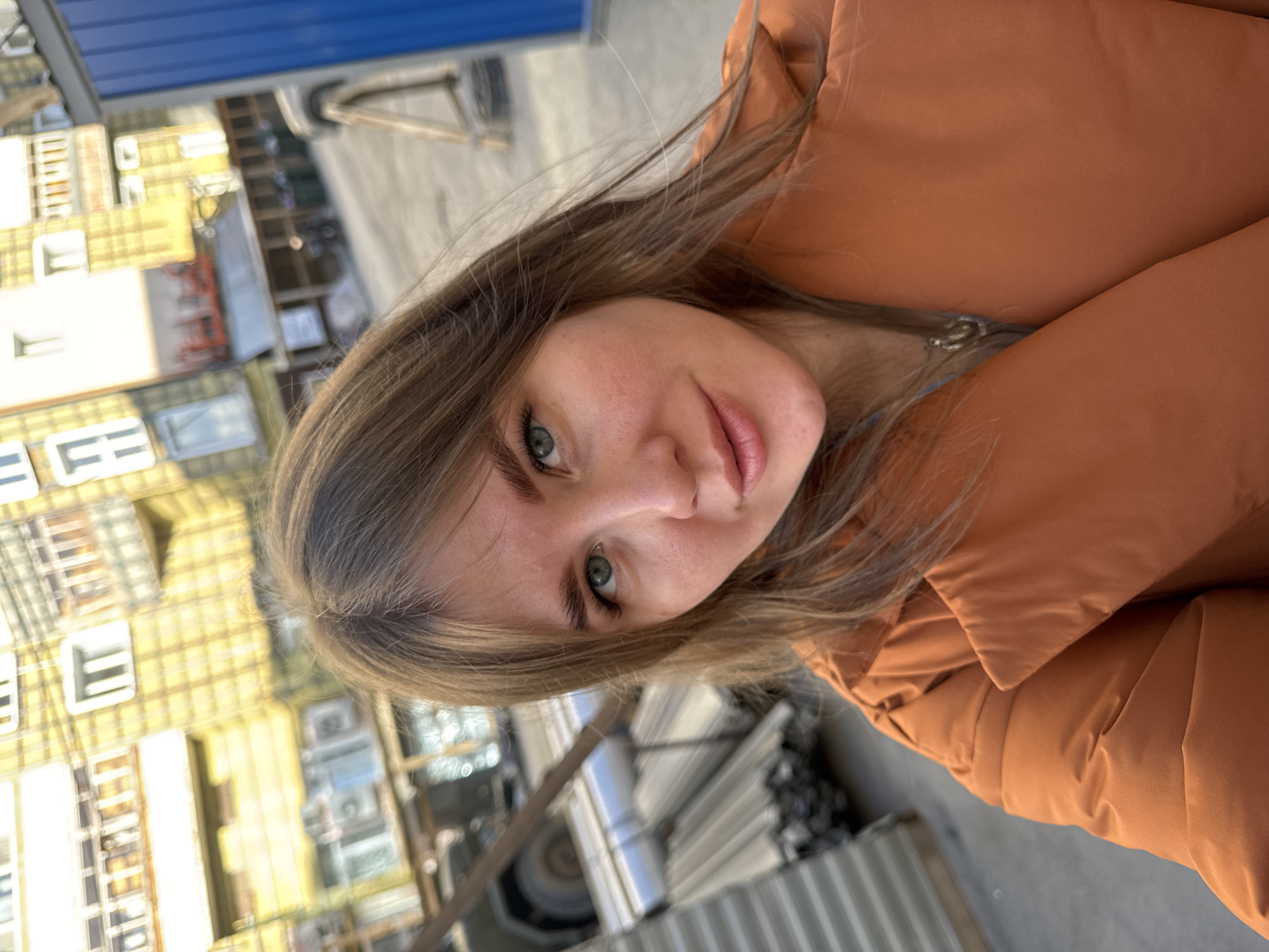 A young woman with long, light brown hair and green eyes is photographed in a slightly angled close-up shot. She is wearing a burnt orange puffer jacket. The background features an urban setting. Structures resembling buildings are visible to the left, along with what appears to be construction materials and equipment in the background. The presence of building elements indicates a location where construction or renovation is taking place.
Although the location is stated as Perm, Russia, there is no definitive evidence within the image itself to confirm this. The image primarily focuses on the woman and the immediately surrounding area, offering little contextual information to identify the specific geographic location definitively.