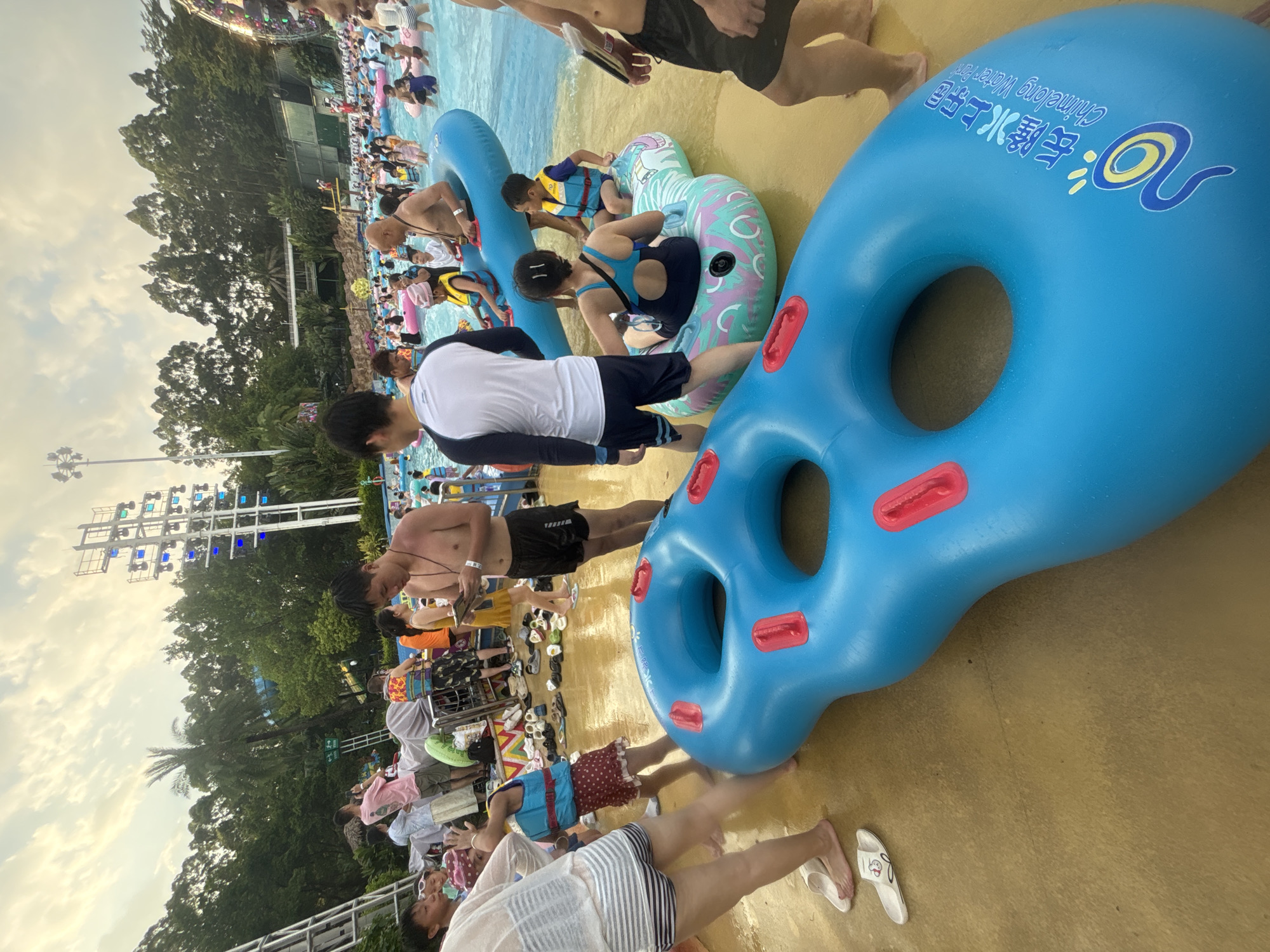 The image depicts a crowded scene at a water park in Panyu District, China. In the foreground, a large, bright blue inflatable tube with red handles lies on a wet, sandy-colored surface. The tube has Chinese characters and a logo on it, identifying it as being from "Chimeelong Water Park." Several people are visible around the tube and in the background.
To the left, a person wearing a white and grey striped top and striped shorts is standing with their back to the camera, partially obscured by another inflatable. Next to them, a young child in a red polka-dot skirt and a blue life vest is facing away from the camera. Further back, a man in a pink shirt and shorts is holding a phone up as if taking a picture.
In the middle ground, a large pool filled with people is visible. Many individuals are floating on various inflatable rings, and some are standing in the shallow water. A man in a white and blue long-sleeved shirt and dark shorts is facing the pool, looking down. A woman in a blue swimsuit is sitting on a colorful inflatable, facing away from the camera, with a young boy in a yellow life vest beside her on the same inflatable.
The background shows lush green trees and a large structure with multiple colored lights, likely a waterslide or other attraction. The sky above is cloudy with patches of light, suggesting it is either early morning or late afternoon. The overall impression is one of a busy recreational environment.