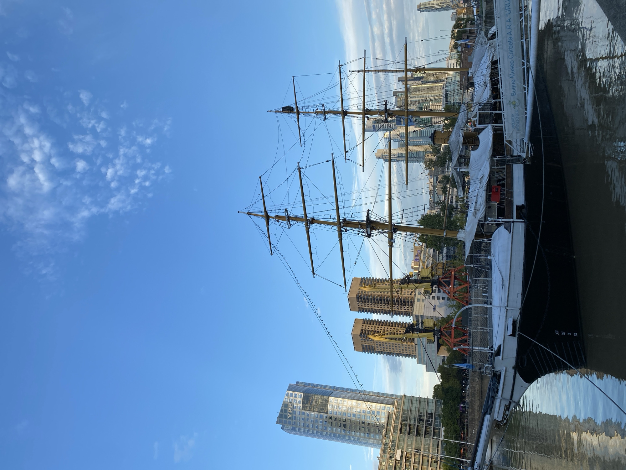 The image shows the "Buque Museo Corbeta A.R.A. Uruguay," a historic sailing ship docked in Buenos Aires, Argentina. The ship has tall masts and rigging. The hull is black, and part of the ship is covered with white canvas or tarps. Modern buildings, including high-rise apartments and hotels, are visible in the background. The sky is blue with scattered clouds. The water reflects the ship and sky. There are no people visible in the frame.