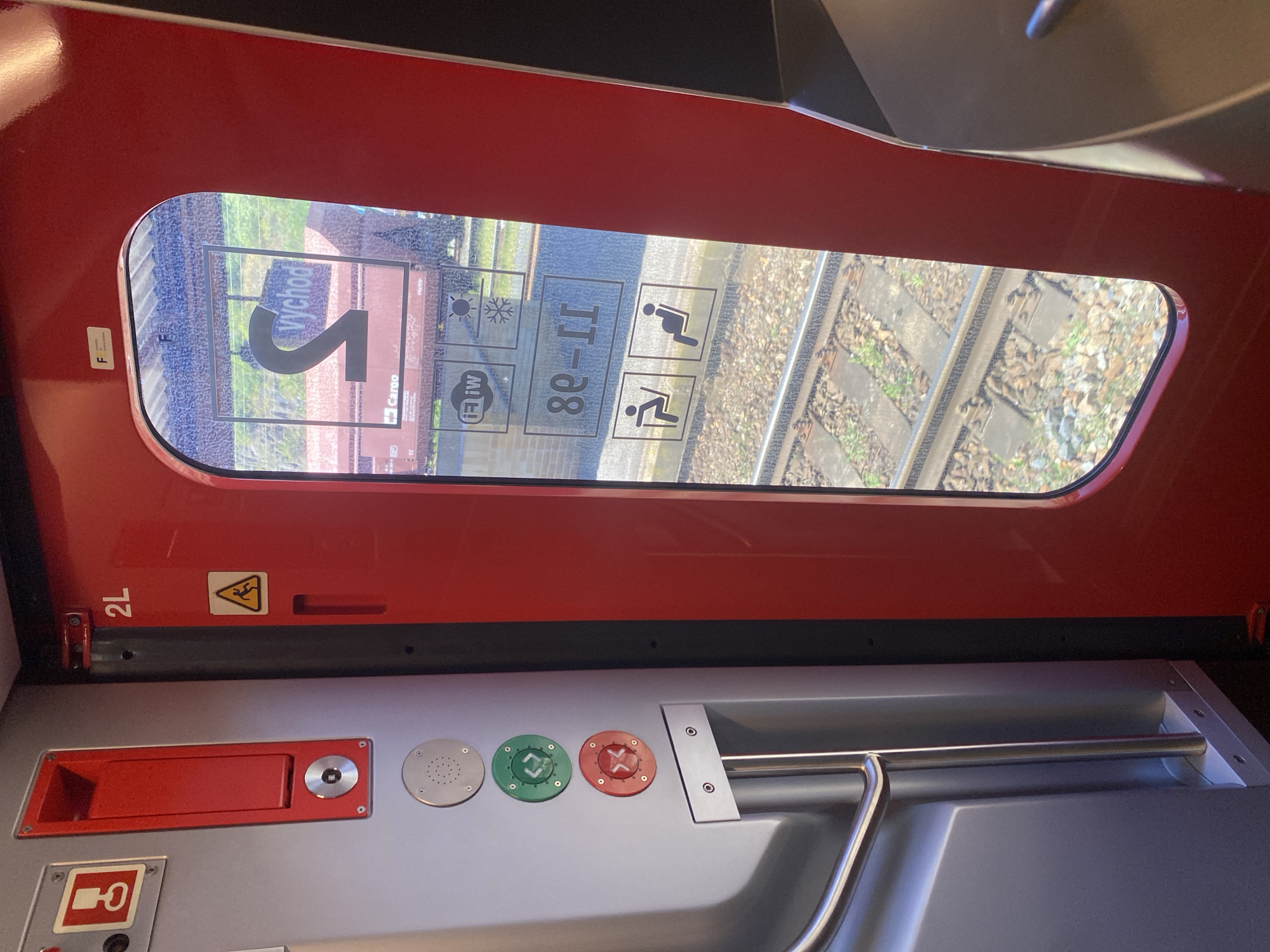 This image captures the interior of a train car at the Bílina train station in the Czech Republic. The focus is on the red train door with a large window that looks out onto the train tracks and a platform. Inside the train car, to the left of the door, a gray panel features a red recessed handle and several circular buttons. One button is red with a white symbol of an 'X', another is green with a symbol of arrows indicating movement in opposite directions, and a third circular component appears to be a speaker. Above these buttons, a red compartment with a handle is visible.
Through the train door window, the word "Východ" (meaning "Exit" in Czech) is displayed in large black lettering on a white background, superimposed over an image of train cars. Below this, there are several smaller icons: one depicting Wi-Fi signal strength, another showing a sun and a snowflake, and two icons illustrating seating arrangements, one with a person in a reclining position and another with a pregnant person. The exterior view through the window shows train tracks and ballast. The overall scene suggests a functional environment within a public transportation vehicle.