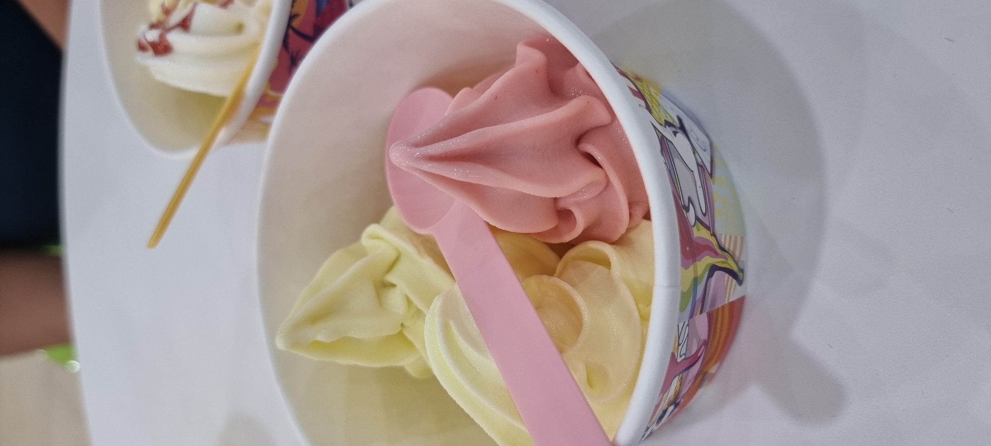 An outdoor scene captures a close-up of two paper cups filled with soft-serve ice cream. The cup in the foreground is white with a colorful unicorn design around its base, containing two swirls of pale yellow ice cream and one swirl of pink ice cream. A pink plastic spoon rests on top of the ice cream. A second white cup, partially visible behind the first, contains white ice cream topped with red syrup or sauce and slivered nuts, with a yellow stick protruding from it. The cups are placed on a white surface, possibly a table, with a blurred background showing a person's arm. The scene suggests a casual outdoor setting, likely in a park or recreational area, given the presence of ice cream and the bright, natural lighting. The location in the city of Aktobe, Kazakhstan, is not visually indicated by any landmarks or environmental cues in the image.