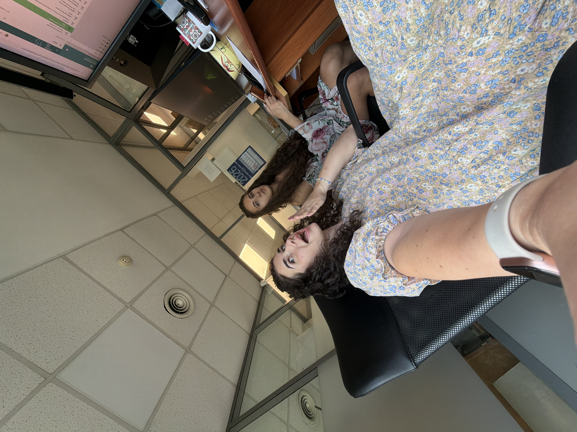Two women are in an office environment, which is likely located in Sofia, Bulgaria, as suggested by the presence of a calendar with "2026" visible. The woman in the foreground is seated in a black office chair and is wearing a light-colored dress with a floral pattern. She is holding a phone and appears to be taking a selfie, with her face looking towards the camera with a surprised expression. Her left arm, wearing a white watch, is extended. Behind her, a second woman is seated at a desk, also wearing a floral dress. She is looking towards the camera and holding a phone. The office has a suspended ceiling with fluorescent lighting and air vents. The desk has two computer monitors, a mug with a colorful pattern, and papers. The background includes glass partitions, suggesting a modern office layout.