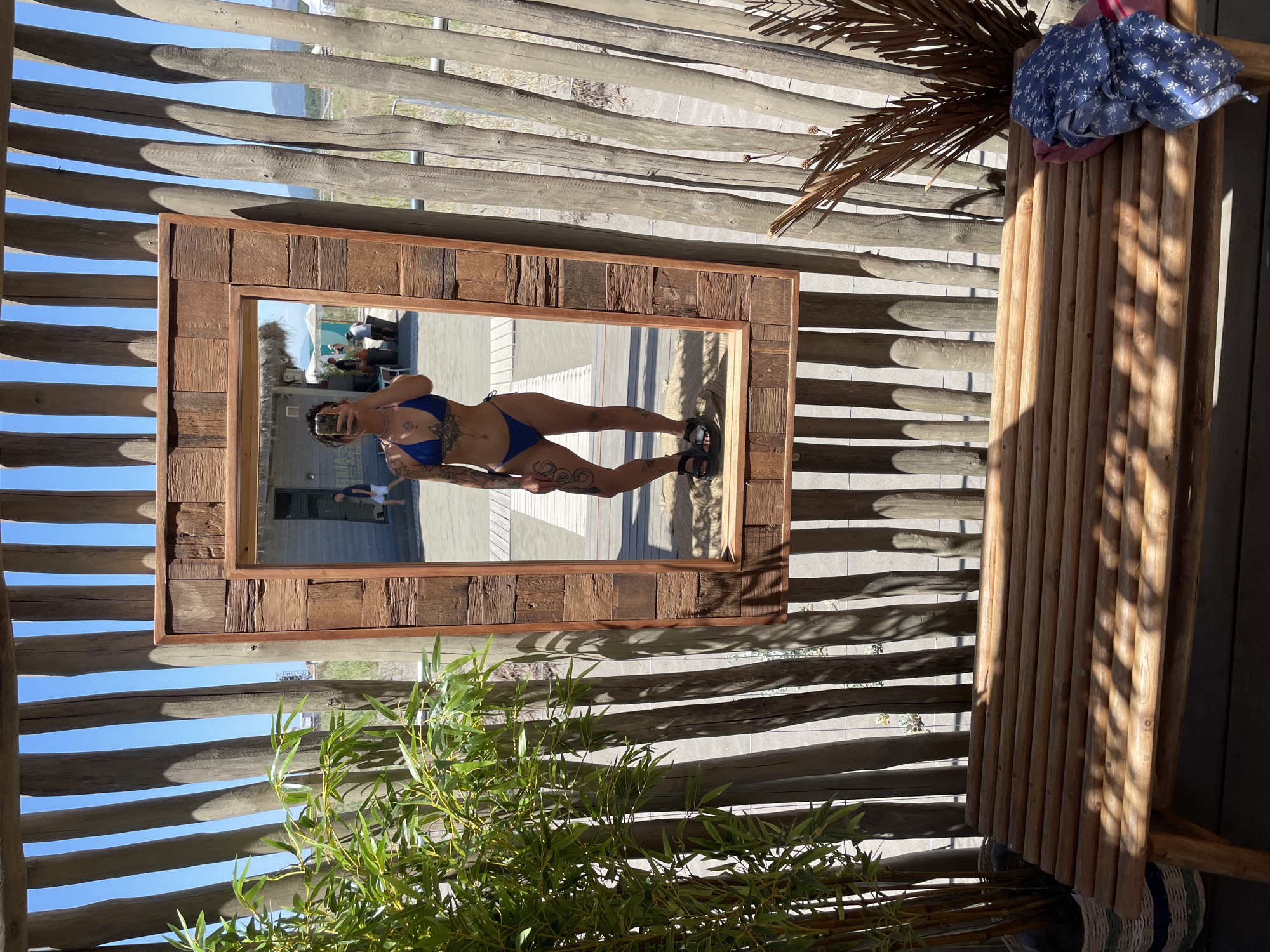 The image depicts a woman taking a mirror selfie in a rustic setting. She is wearing a blue bikini and has tattoos on her arms and legs. The mirror is framed with weathered wood and hangs on a structure made of vertical wooden poles. In the reflection, other people and beach umbrellas can be seen in the background, suggesting a beach or outdoor recreational area. In the foreground, there is a wooden bench with some folded clothing on it and a potted plant to the left. The overall environment suggests a sunny, outdoor location, possibly a beach resort in Donji Štoj, Montenegro.