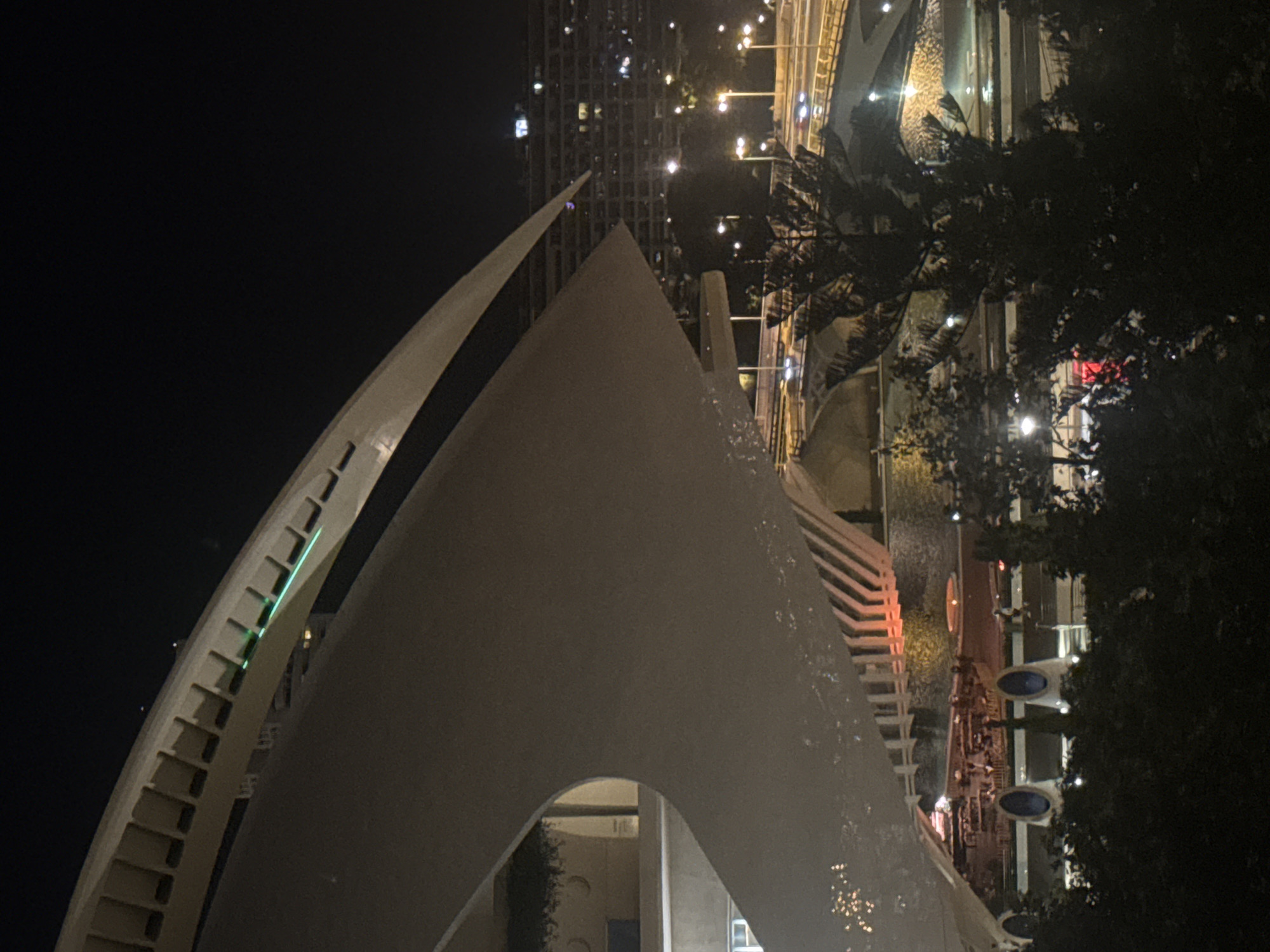The image shows a night view of the City of Arts and Sciences in Valencia, Spain, featuring the Hemisfèric. A large, white, curved architectural structure dominates the foreground, casting shadows. In the background, a multi-story building with illuminated windows is visible. Below the main structure, a body of water reflects lights from the surroundings, and bridges with traffic are present. Trees and foliage partially obscure the lower part of the scene. The overall atmosphere is dark, with artificial lighting creating highlights and illuminating the modern architecture.