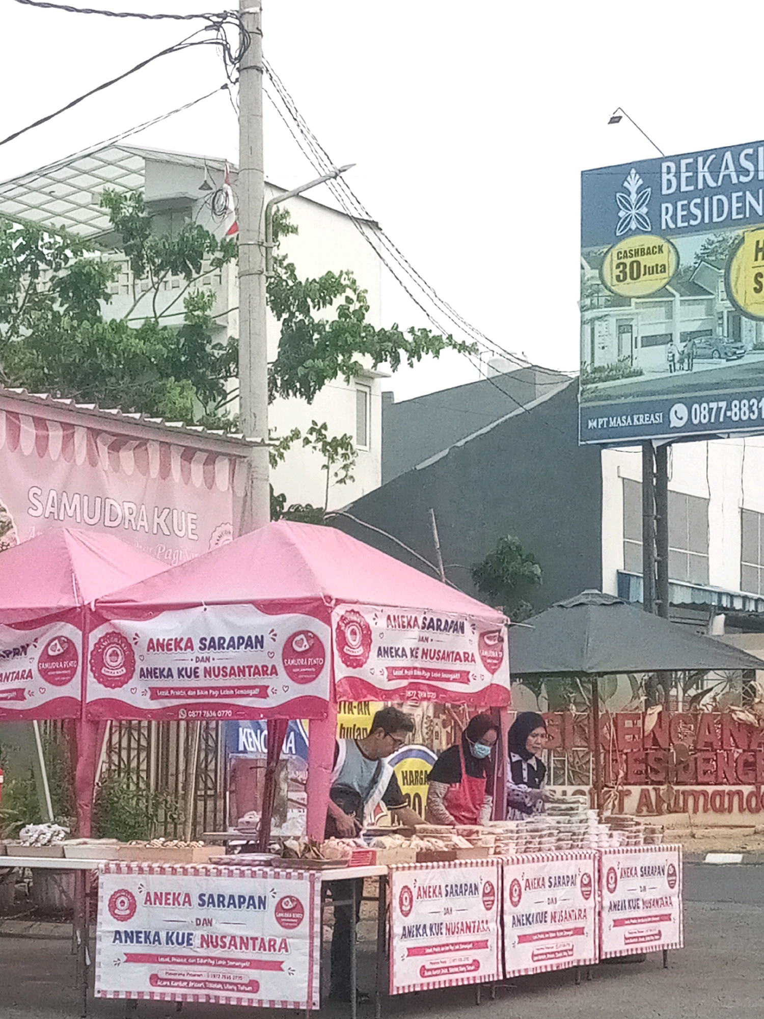 In Bekasi, Indonesia, a food stall is set up on the roadside, featuring pink tents with white banners. The banners advertise "ANEKA SARAPAN DAN ANEKA KUE NUSANTARA," indicating a variety of breakfast items and traditional Indonesian cakes. Several food containers are arranged on display tables. Three individuals are visible behind the stall: one man in a blue shirt and red apron, and two women wearing hijabs; one of them is also wearing a face mask. In the background, a billboard advertises "BEKASI RESIDENCE," with a phone number and "PT MASA KREASI" visible. Utility poles and wires are present, along with trees and buildings. The overall scene depicts an outdoor commercial activity.