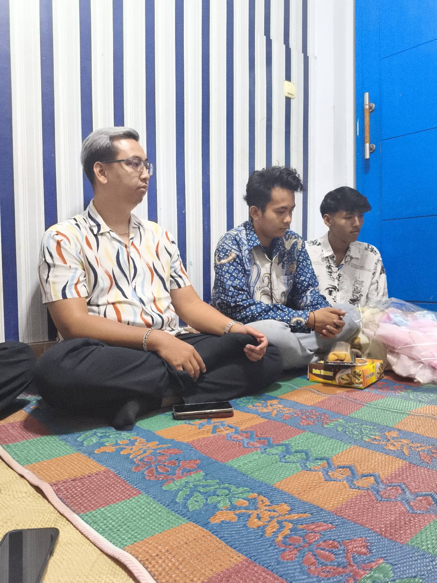 Three young men are seated on a woven mat in what appears to be a residential setting in Depok, Indonesia. The man on the left has light gray hair and is wearing glasses and a colorful, short-sleeved patterned shirt. He is also wearing black pants and a silver bracelet. To his right, a man in a blue patterned shirt is looking down, and further to the right, a third man in a white patterned shirt is also looking down. In front of them, on the mat, is a box of snacks and a plastic bag containing pink items. The background features a wall with vertical blue and white stripes, and to the right, a bright blue door with a silver handle. The mat itself has a colorful geometric pattern with floral motifs. The lighting suggests an indoor environment.