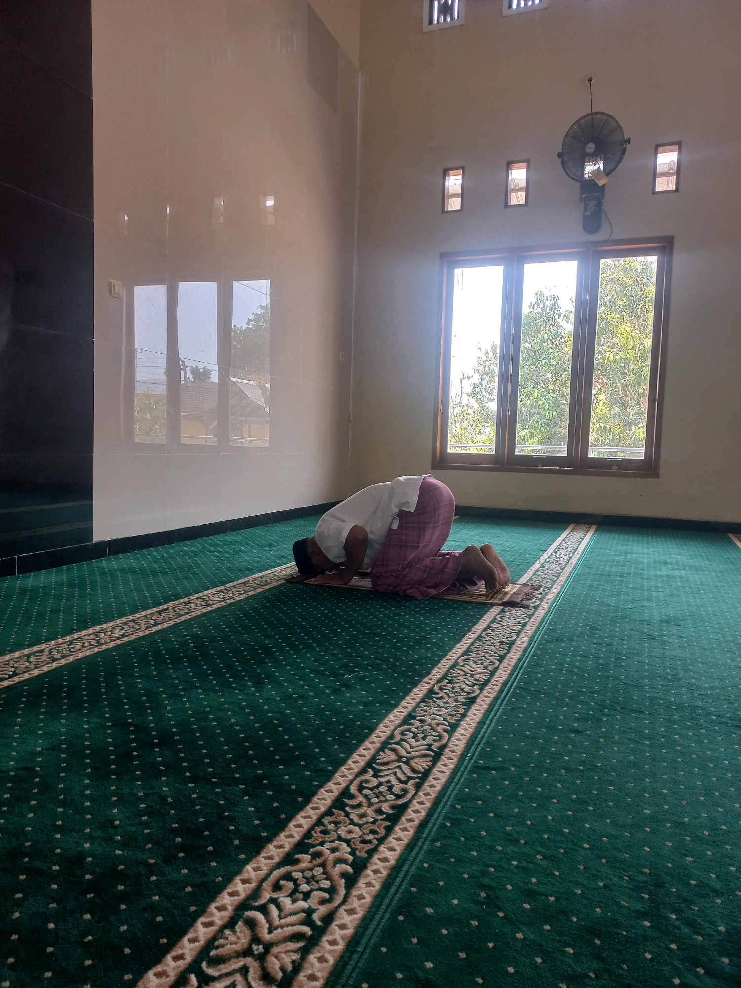 A person is praying on a prayer mat in a mosque. The person is wearing a white shirt and a checkered pink and white sarong. They are kneeling with their forehead touching the mat. The mosque has a green carpet with a decorative border and small dots. There are windows in the background with trees visible outside. A ceiling fan is mounted on the wall. The room appears to be clean and well-lit. This mosque is located in the city of Nguntoronadi, Indonesia.