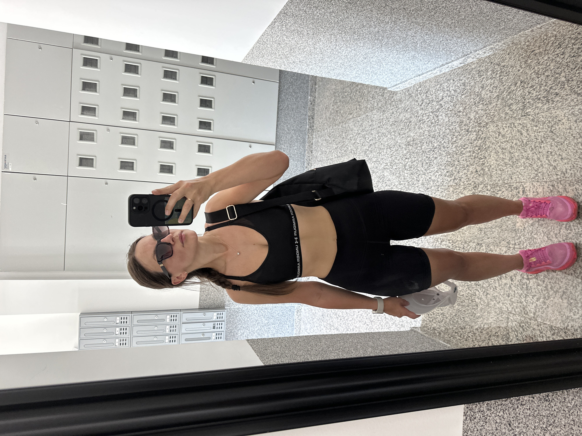 A young woman, with her hair in a braid, stands in a hallway of lockers and takes a mirror selfie. She is wearing a black sports bra, black bike shorts, bright pink athletic shoes, and sunglasses. She holds a white face mask in her left hand and has a black backpack slung over her right shoulder. The lockers are grey and appear to be in a gym or locker room. The floor is speckled grey.