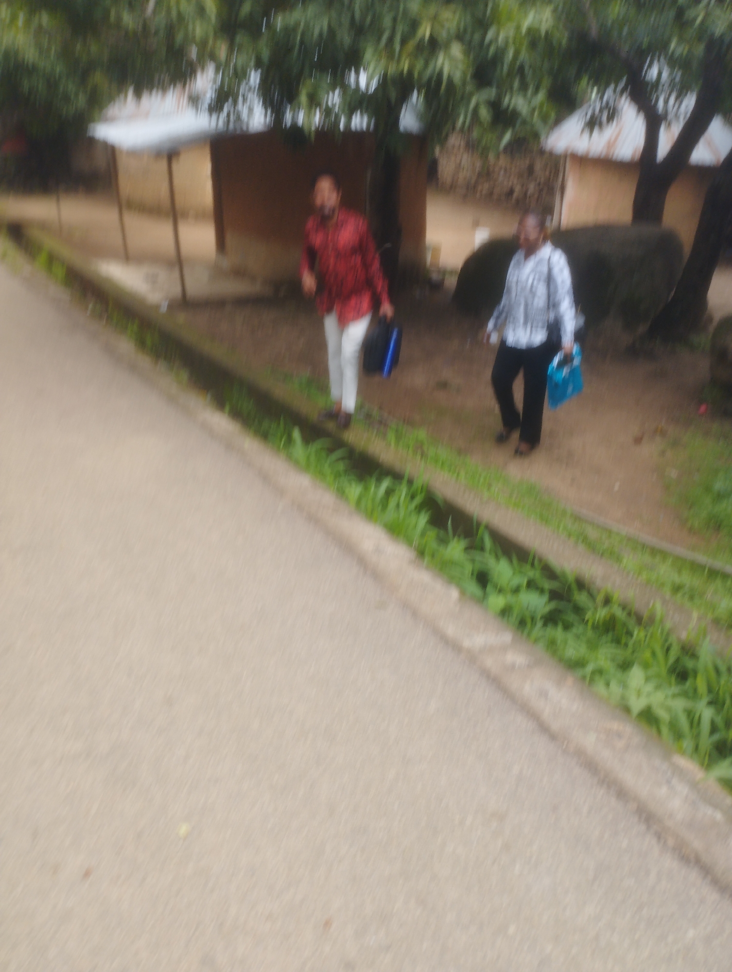 The image shows a street scene in Jos, Nigeria. Two individuals are walking on the side of a road. The person on the left is wearing a red patterned shirt and white pants, carrying a dark blue bag. The person on the right is wearing a checkered shirt and black pants, carrying a blue bag. They are walking past a row of buildings with corrugated metal roofs, and there is vegetation along the side of the road. The road surface appears to be paved.