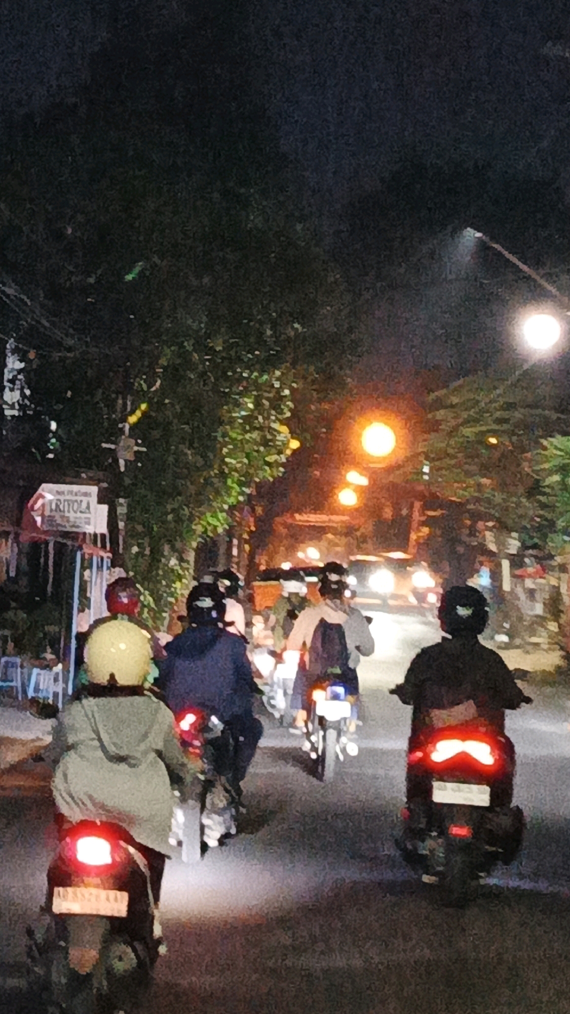 A group of people are riding motorcycles on a street at night in Surakarta, Indonesia. The street is lined with trees and buildings. Several streetlights are visible, casting an orange glow on the scene. Some of the motorcycles have their brake lights illuminated, indicating they are slowing down or stopping. The people on the motorcycles are wearing helmets, and some are wearing casual clothing. The overall impression is a common nighttime scene in a busy urban area.
