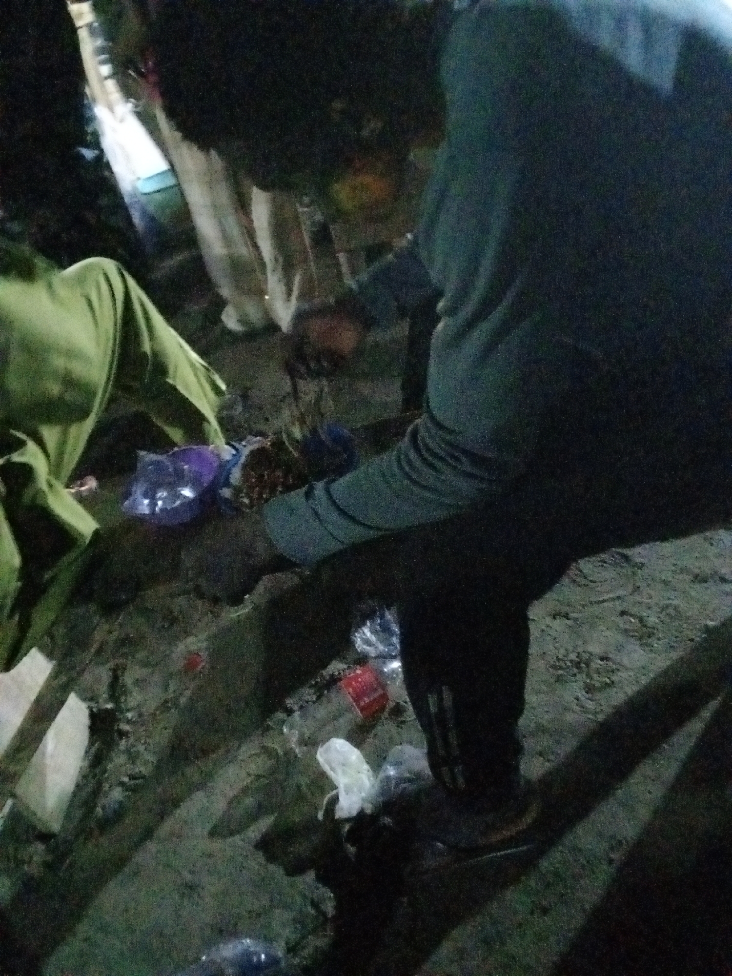 The image shows a person wearing a long-sleeved grey shirt and dark pants with light-colored stripes down the side of the leg. The person is crouched over a collection of items, including plastic bags and food wrappers, lying on the ground, which appears to be dirt. There are other people standing in the background. The setting appears to be outdoors. The location is in the city of Ado, Nigeria, however, the image does not provide specific landmarks or details that would identify the precise area within Ado.