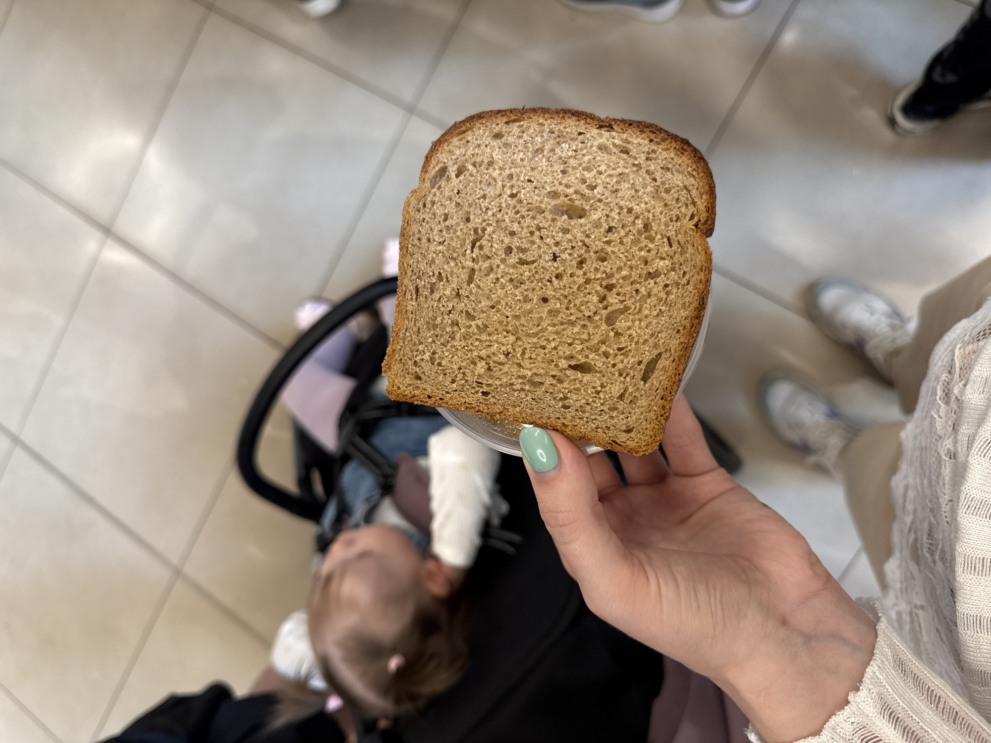 A top-down indoor shot shows a woman holding a slice of brown bread in a clear plastic container. The woman has fair skin and a light green fingernail. A baby in a stroller is visible in the background. The floor is tiled. Other people's legs and feet are also visible in the background. The location cannot be determined.