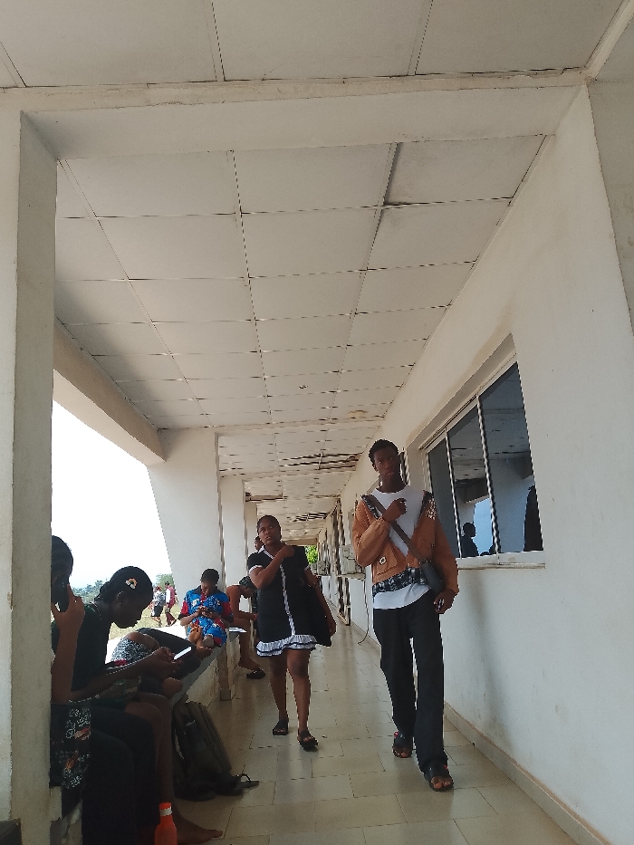 Here is a description of the image:
Location: Covered outdoor corridor in Umueshi, Nigeria.
Environment: Ceiling tiles are visible, with white-painted walls and pillars. Windows are present on the right-hand side of the corridor.
People: Several individuals are present. Two are walking toward the camera. A group of people are seated on a bench to the left, some using phones.
Objects: Bags are on the floor near the seated individuals. Windows are along the right side. A water bottle is on the ground in the lower left.
Actions: People walking and sitting. Some are using phones.
Interactions: People are interacting with their devices. The two individuals walking are headed toward the camera.