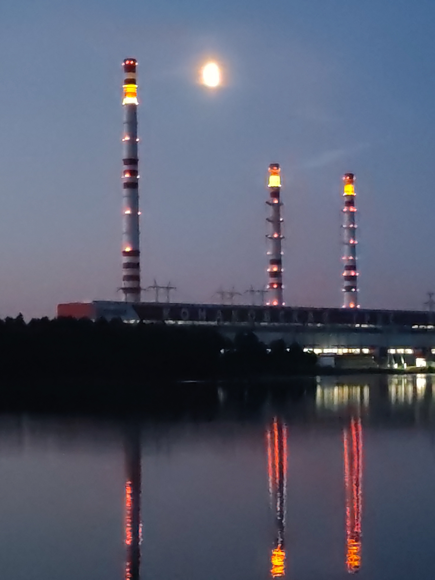 The image shows a power plant with three tall, striped smokestacks at dusk, likely in an industrial area. The smokestacks are white with red bands and illuminated with orange lights at intervals, suggesting they are functional. The tallest smokestack is on the left, followed by two slightly shorter ones to the right. In the background, a large industrial building with lights is visible. The foreground features a body of water, likely a river or lake, reflecting the lights of the smokestacks and the building. A bright, circular object, possibly the moon, is visible in the upper left portion of the sky. The overall scene depicts an industrial landscape at twilight, with the illumination of the power plant contrasting with the darkening sky and water. The location is identified as Unknown, Unknown.