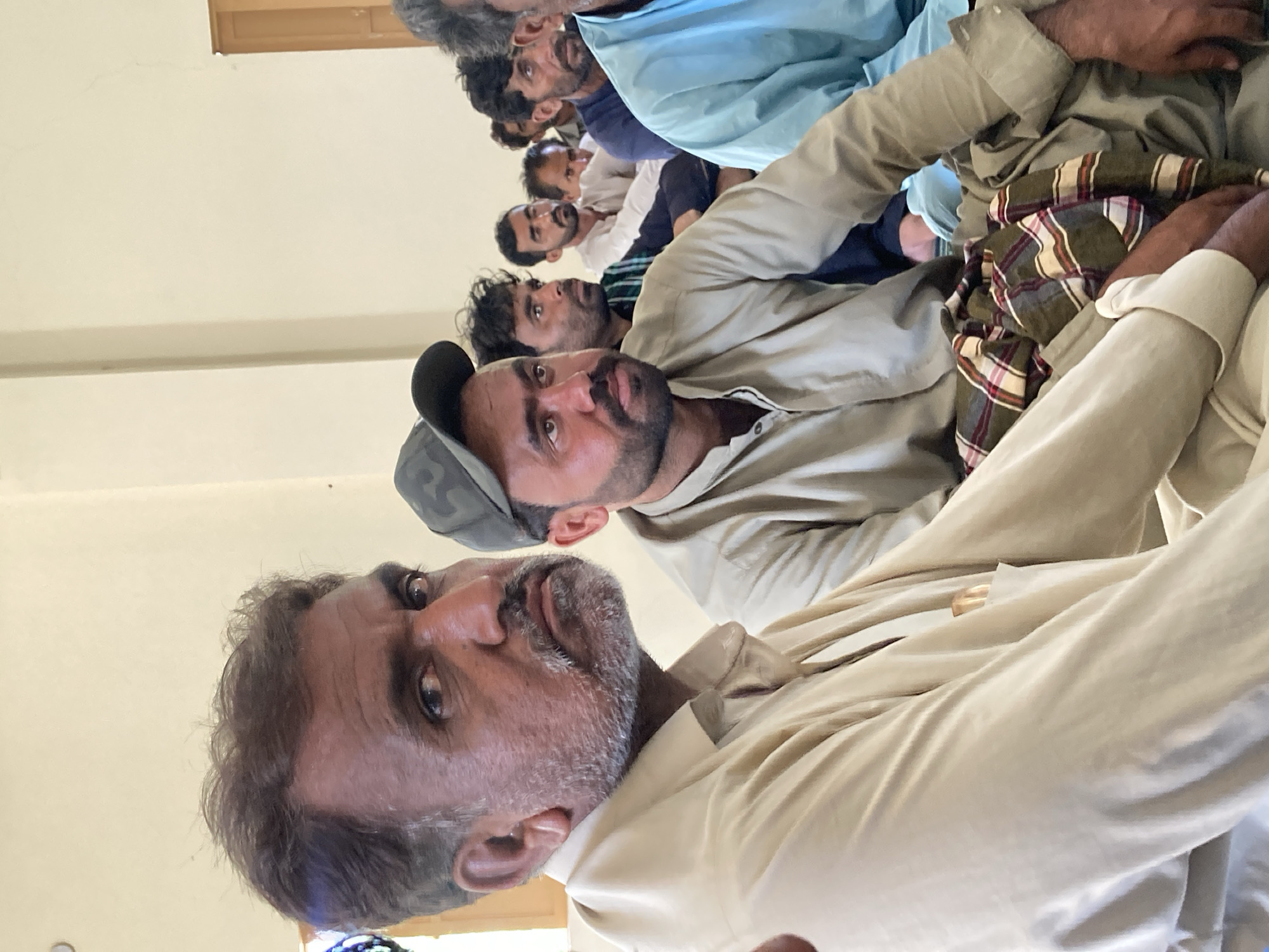 The image shows a group of men sitting in an indoor setting, likely a meeting or gathering. The men are dressed in traditional Pakistani clothing. The location is assumed to be Goth Ali Marād, Pakistan. There are no visible objects except for the clothing worn by the men. Their actions include sitting and looking toward the front. They are sitting close together, implying some interaction or shared purpose. The environment consists of plain, light-colored walls and a door in the background.