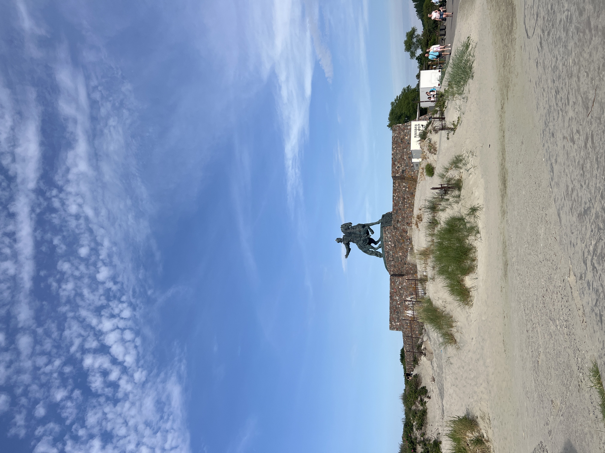 The image captures an outdoor scene on a bright, partly cloudy day. In the foreground, a sandy embankment with sparse green vegetation rises towards a large stone structure. Perched atop this structure is a dark bronze equestrian statue of a man in historical attire, his right arm extended forward. The statue is set against a backdrop of a blue sky dotted with scattered white clouds.
Behind the statue, a low, rough-hewn stone wall forms the base of the monument. To the right of the statue, a paved walkway leads away from the sandy area. Several individuals are visible on this walkway, some walking and others standing near small white structures, one of which appears to have text on it. The environment suggests a coastal or elevated park setting. This monument is the Peter the Great Monument located in Baltiysk, Russia, marking the westernmost point of the country.
