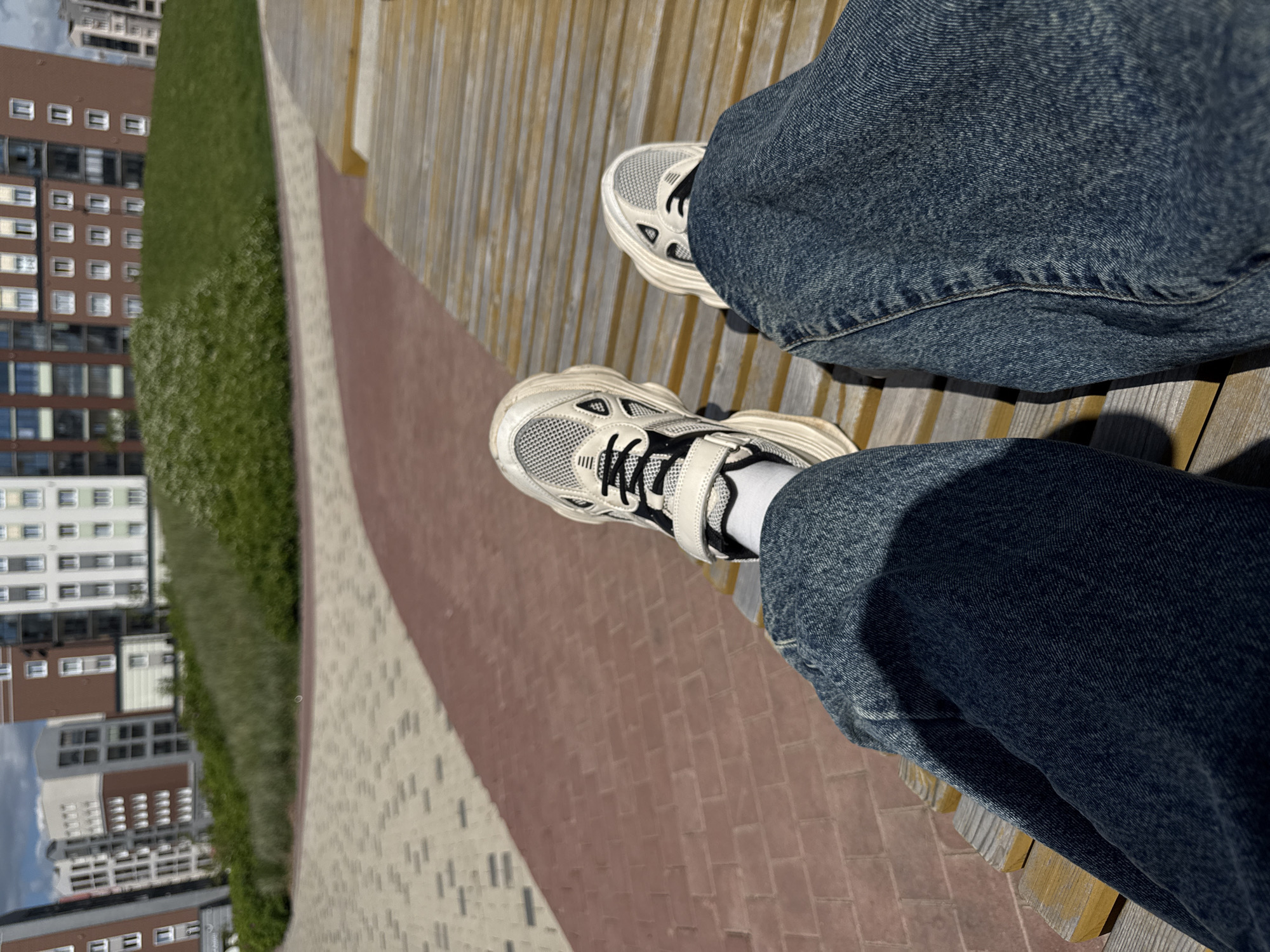 The image presents a high-angle, Dutch-tilted view of a person's legs and feet, likely taken from their own perspective while sitting outdoors. The person is wearing blue denim jeans and white athletic socks, paired with chunky white and light-grey sneakers featuring mesh panels and black laces. Their feet are extended forward, resting on or near a slatted wooden bench or boardwalk.
The scene is an outdoor urban or suburban environment in Pervomaysky, Russia, characterized by clear, sunny weather, as evidenced by the sharp shadows cast by the legs and shoes onto the wooden surface. Directly in front of the person, a distinctive pathway is visible, composed of reddish-brown brick pavers and lighter grey or beige rectangular pavers, arranged in contrasting sections. To the left of the pathway is a vibrant green grassy area, suggesting a park, lawn, or well-maintained green space.
In the background, beyond the green area, several multi-story residential buildings are visible. These structures feature numerous windows, some reflecting the bright sky. The overall impression is one of a relaxed moment spent in a well-developed urban landscape, possibly a public park or a promenade within a residential complex. No discernible text is visible in the image.